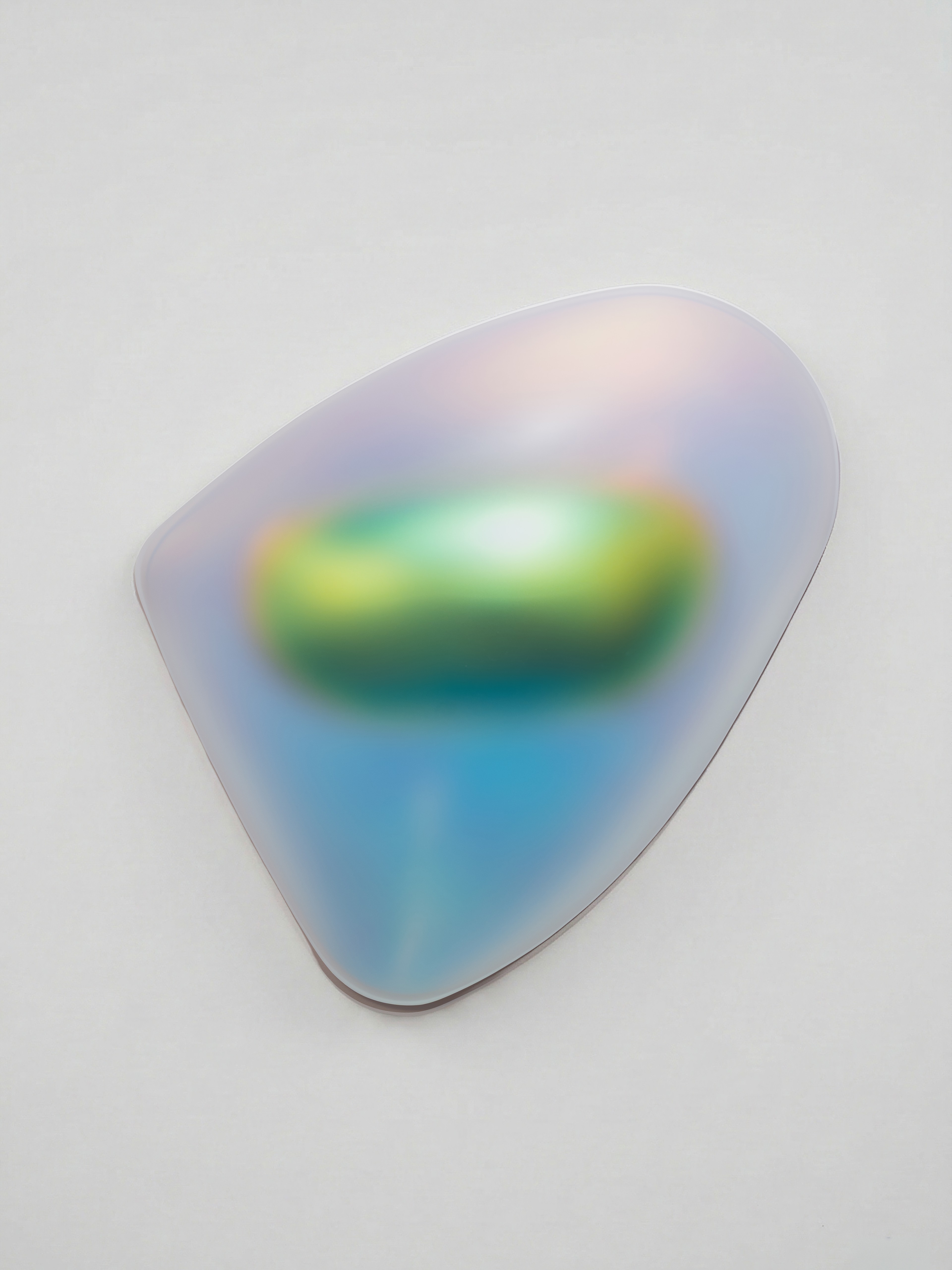 An abstract wall sculpture with a smooth, organic shape resembling a rounded triangle. The surface has an iridescent, translucent finish that shifts in color from green and yellow at the center to blue, violet, and soft pink toward the edges. The artwork appears to glow subtly against a white gallery wall, creating a gentle shadow beneath it.