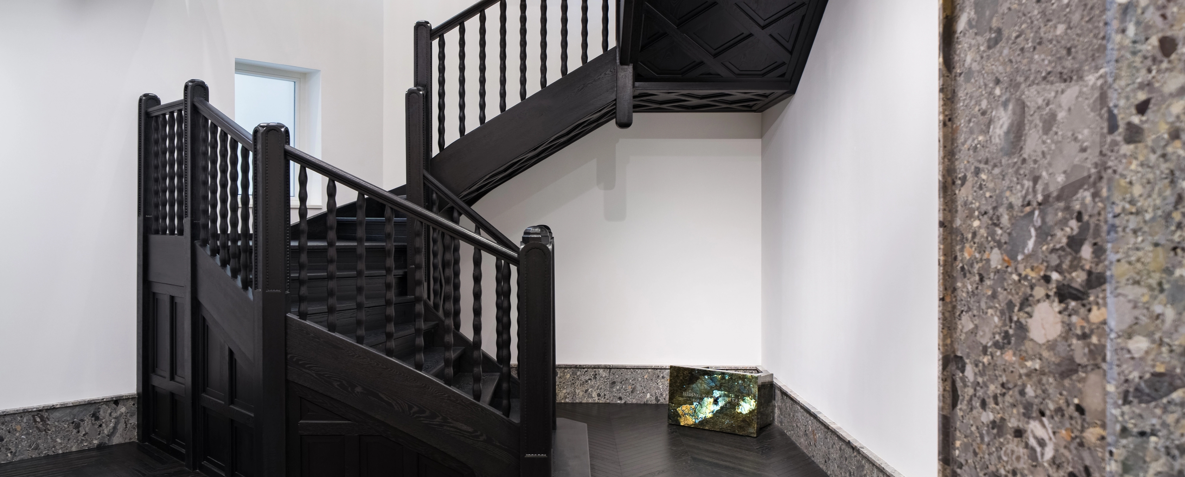 A central staircase with dark wooden balusters and a sculpted ceiling detail, set against white walls. At the foot of the stairs sits a polished green-and-gold stone sculpture with text etched on its surface. The dark herringbone floors and stone baseboard create a refined, modern atmosphere.