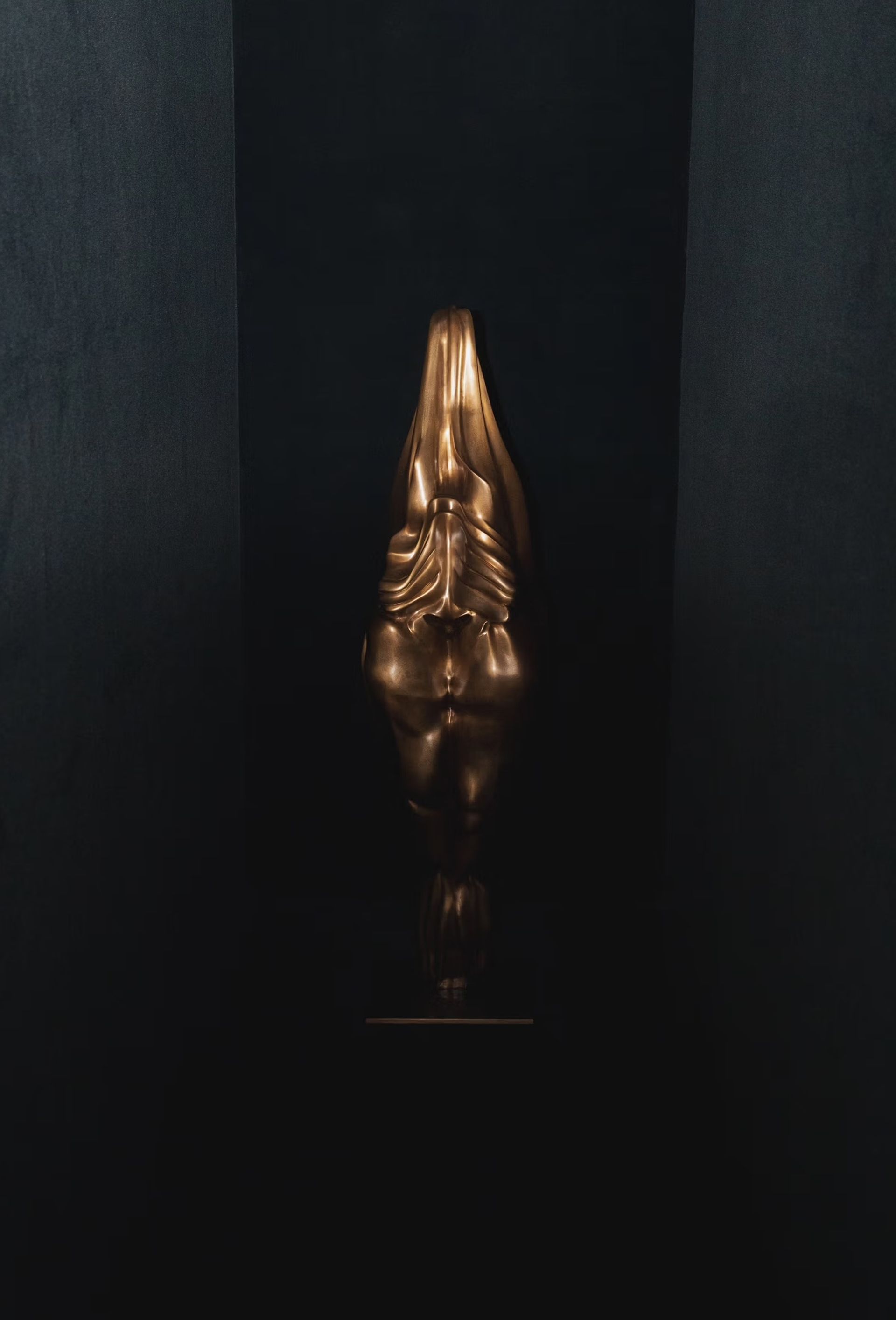 A dimly lit, narrow black corridor containing a tall, elongated gold sculpture that resembles a stylized, draped human torso. The sculpture stands upright against the back wall, its metallic surface catching a subtle light and creating dramatic shadows.