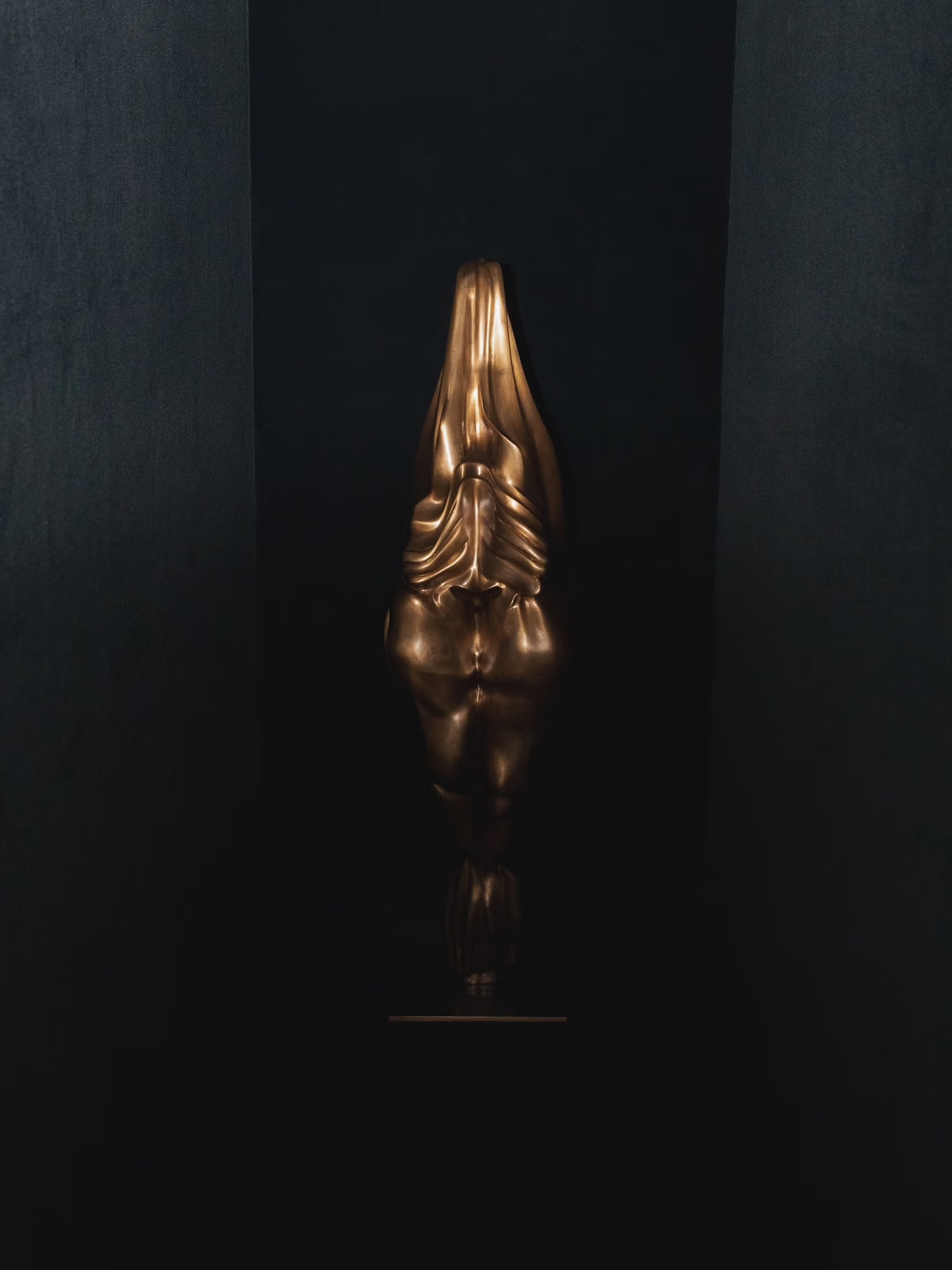 A dimly lit, narrow black corridor containing a tall, elongated gold sculpture that resembles a stylized, draped human torso. The sculpture stands upright against the back wall, its metallic surface catching a subtle light and creating dramatic shadows.