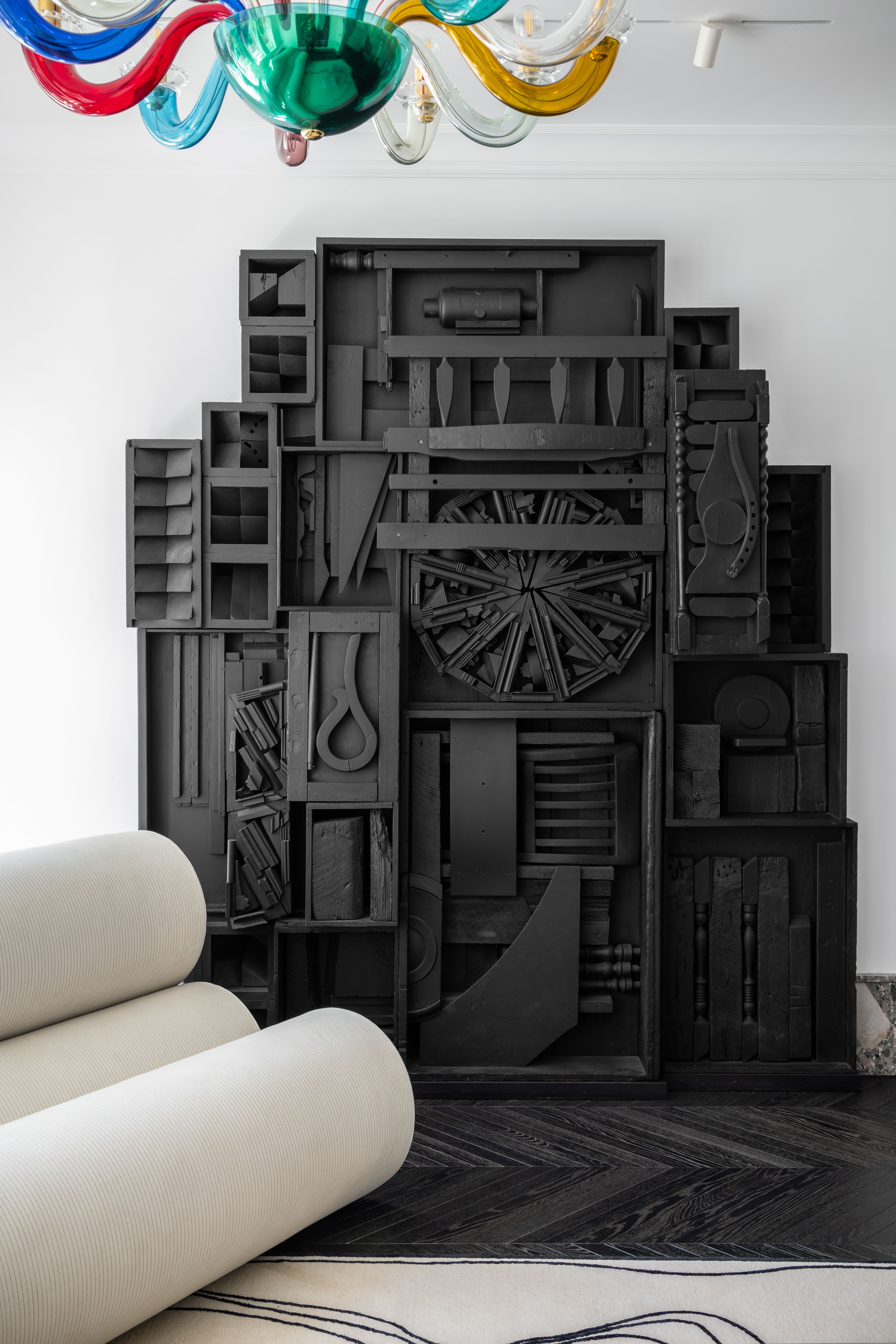 A large black sculptural wall relief composed of assorted wooden elements such as boxes, wheels, and carved forms, painted matte black. In the foreground, a curved white sofa and a colorful glass chandelier add contrast to the monochrome assemblage.