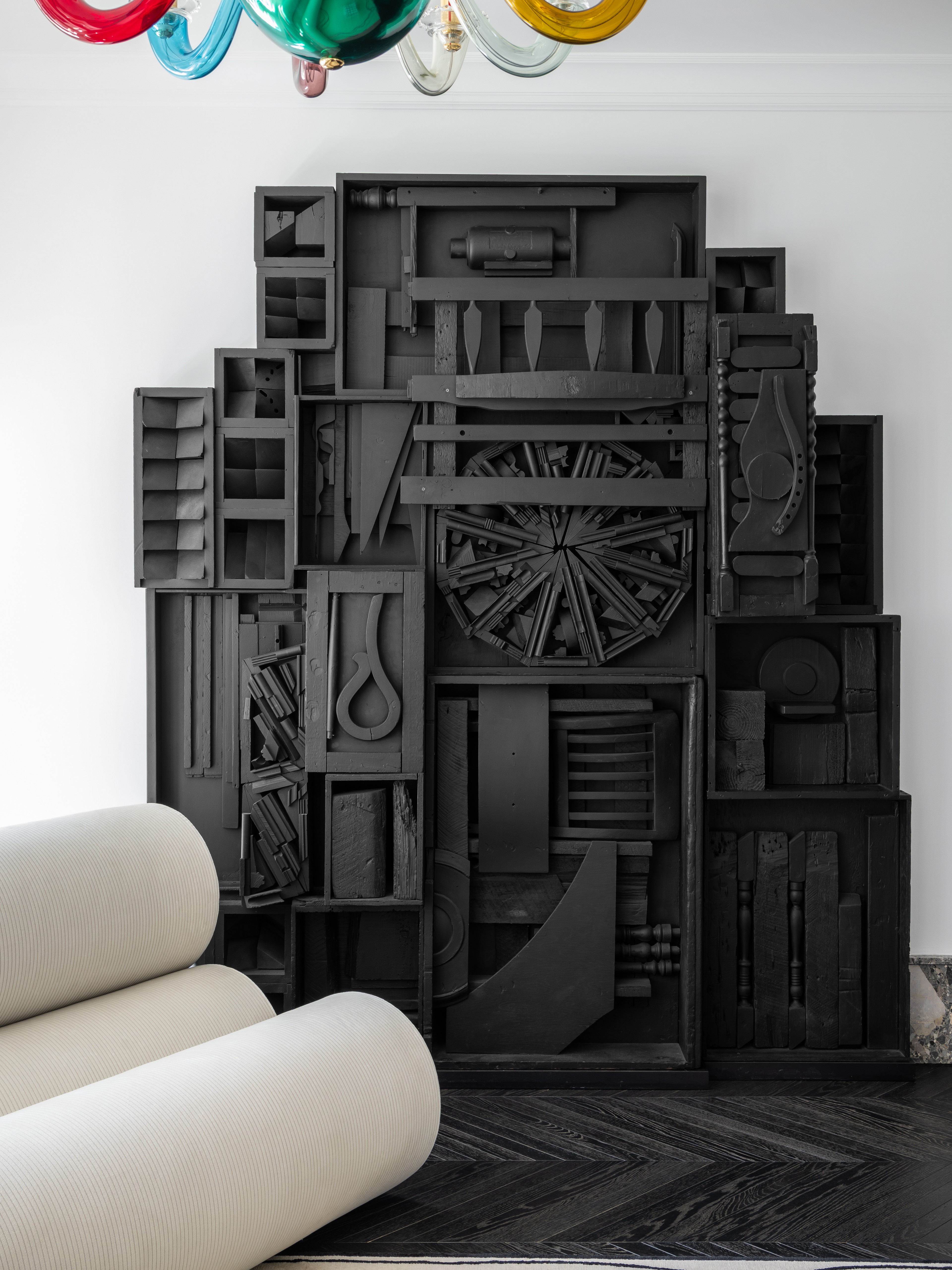 A large black sculptural wall relief composed of assorted wooden elements such as boxes, wheels, and carved forms, painted matte black. In the foreground, a curved white sofa and a colorful glass chandelier add contrast to the monochrome assemblage.