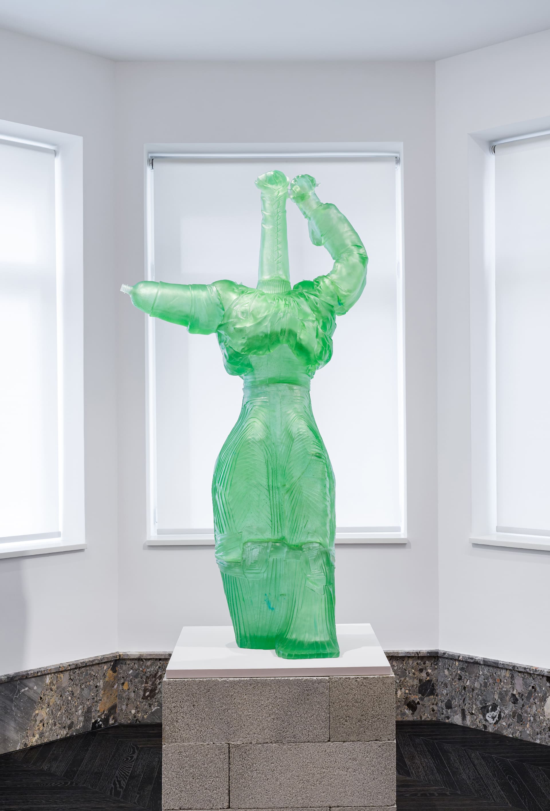 A translucent green sculpture resembling a stylized human figure with raised arms, displayed on a white pedestal made of concrete blocks in a minimalist gallery space with white walls and large windows.