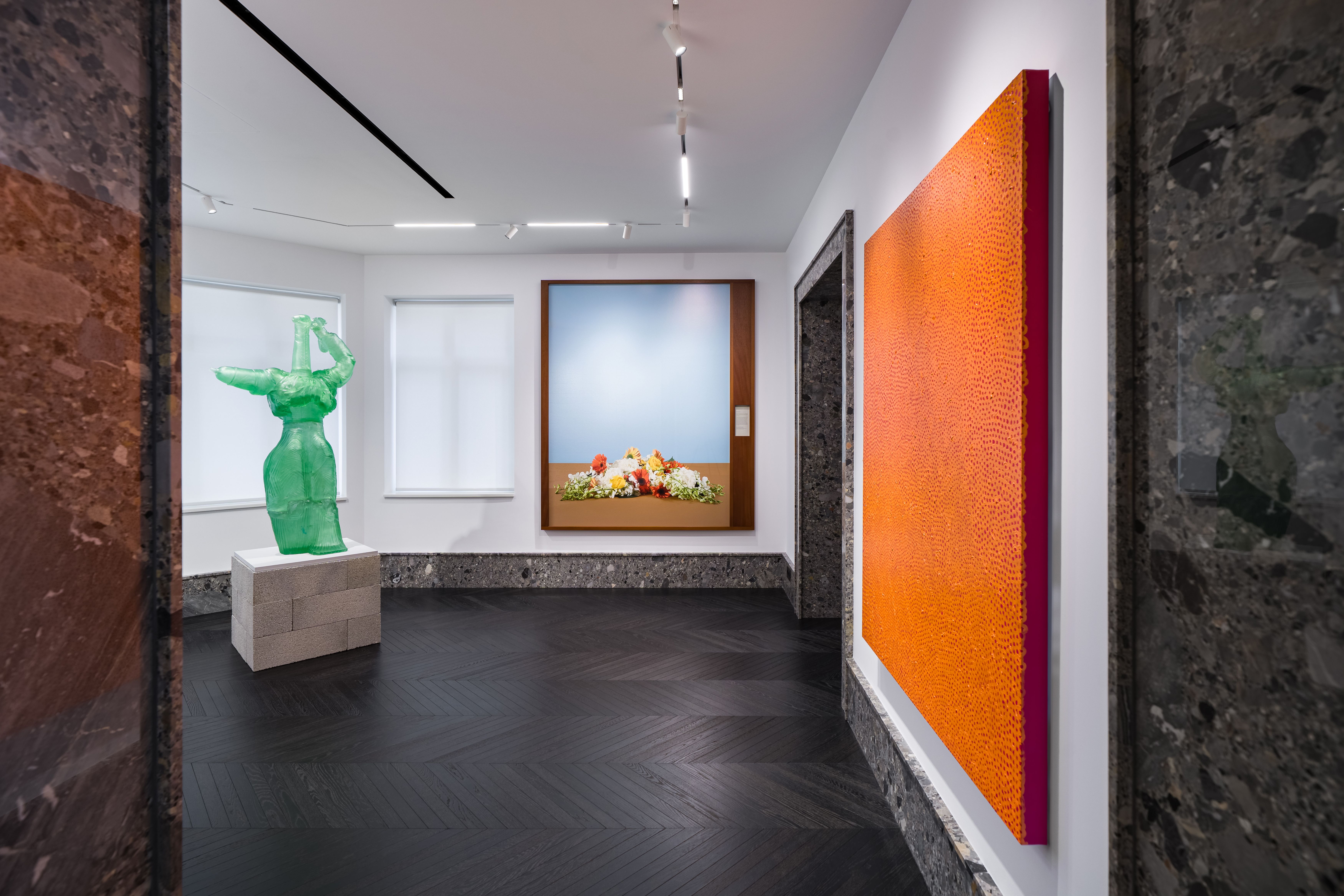 A bright gallery room with an orange textured painting, a translucent green sculpture of a stylized human form, and a framed photograph of flowers against a pale blue background. The space combines modern lighting and dark wood flooring with stone trim around doorways and baseboards.
