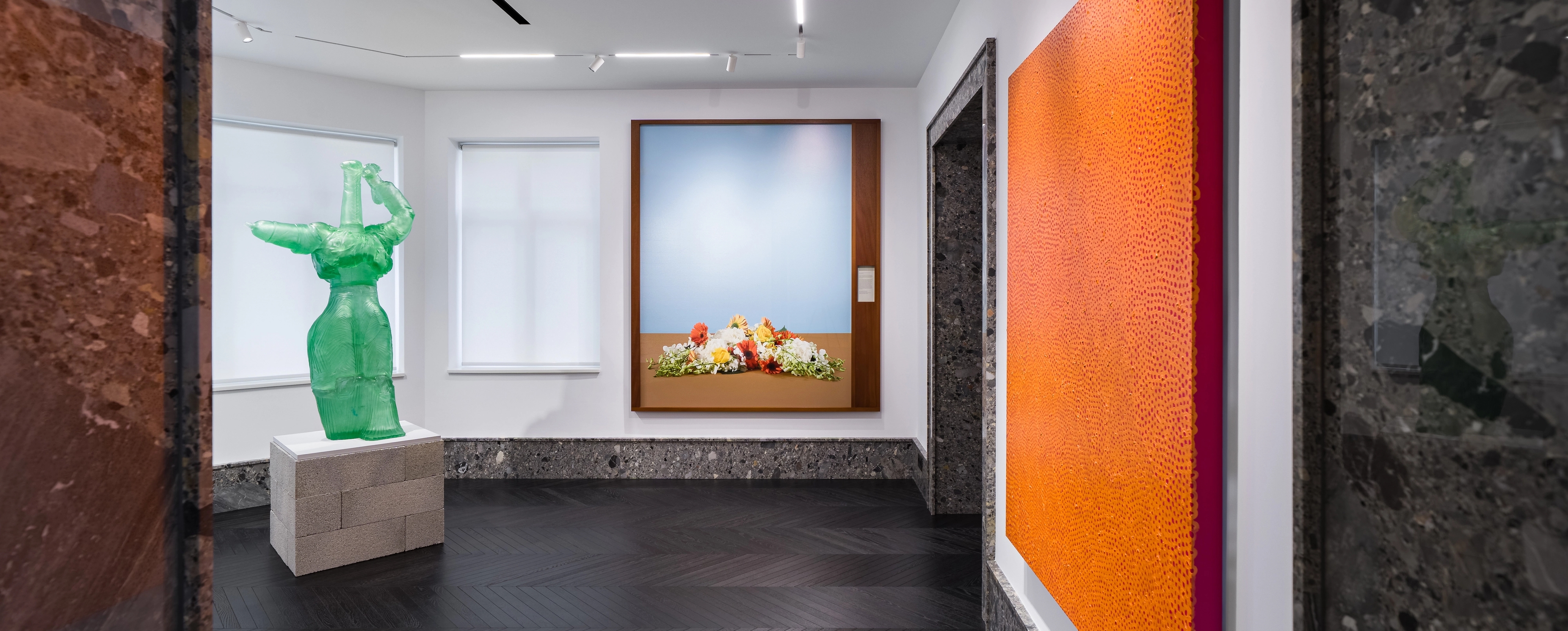 A bright gallery room with an orange textured painting, a translucent green sculpture of a stylized human form, and a framed photograph of flowers against a pale blue background. The space combines modern lighting and dark wood flooring with stone trim around doorways and baseboards.