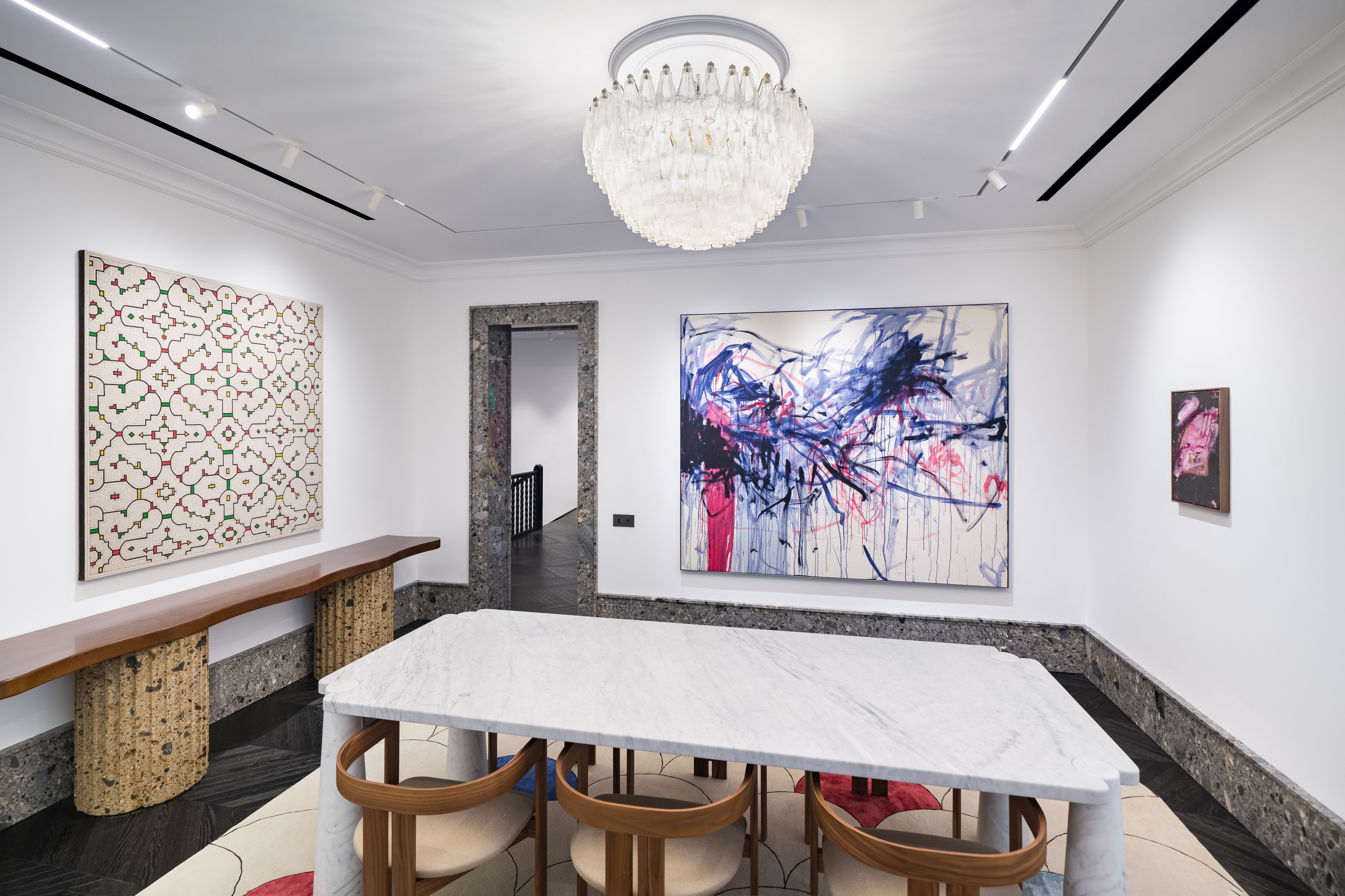 A bright, elegant gallery room featuring two large paintings—one with a linear red and green pattern, another with dynamic blue and red brushstrokes. Curved wooden furniture and a marble table complement the dark herringbone floor and textured stone door frames.