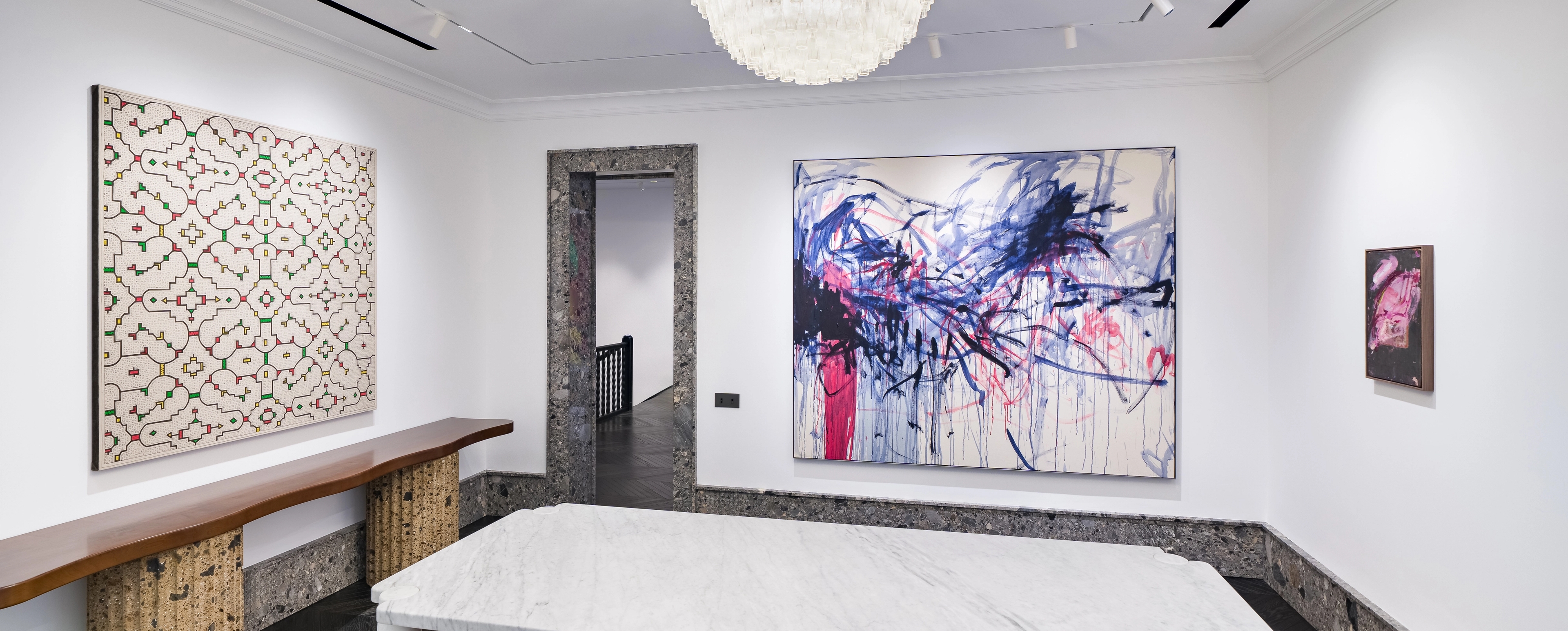 A bright, elegant gallery room featuring two large paintings—one with a linear red and green pattern, another with dynamic blue and red brushstrokes. Curved wooden furniture and a marble table complement the dark herringbone floor and textured stone door frames.