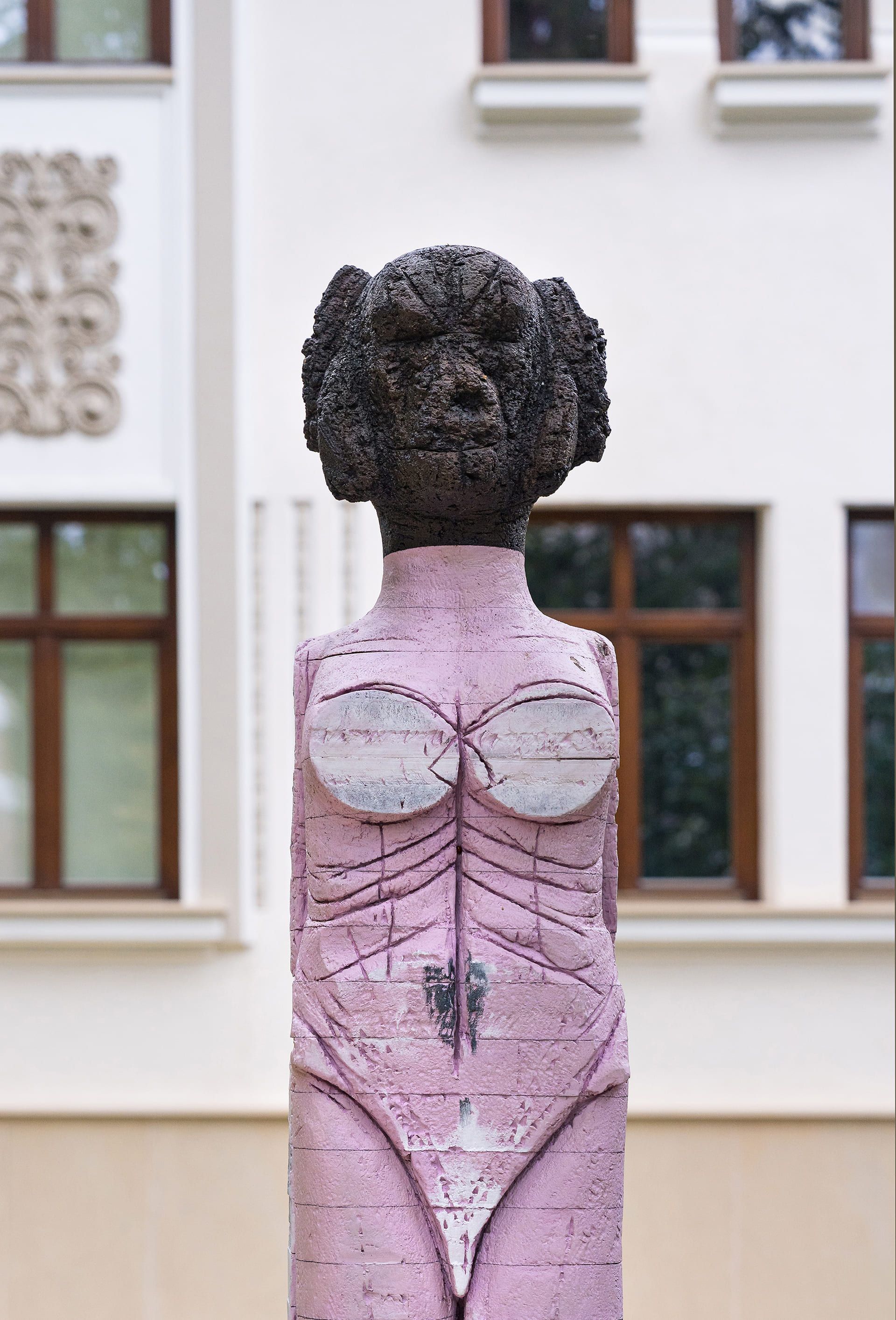 Outdoor sculpture of a stylized human figure with a rough, dark textured head and lower legs. The torso and upper legs are painted a muted pink, with carved linear markings suggesting anatomical features. The chest area features two rounded white forms, and the surface shows signs of weathering and cracks throughout.