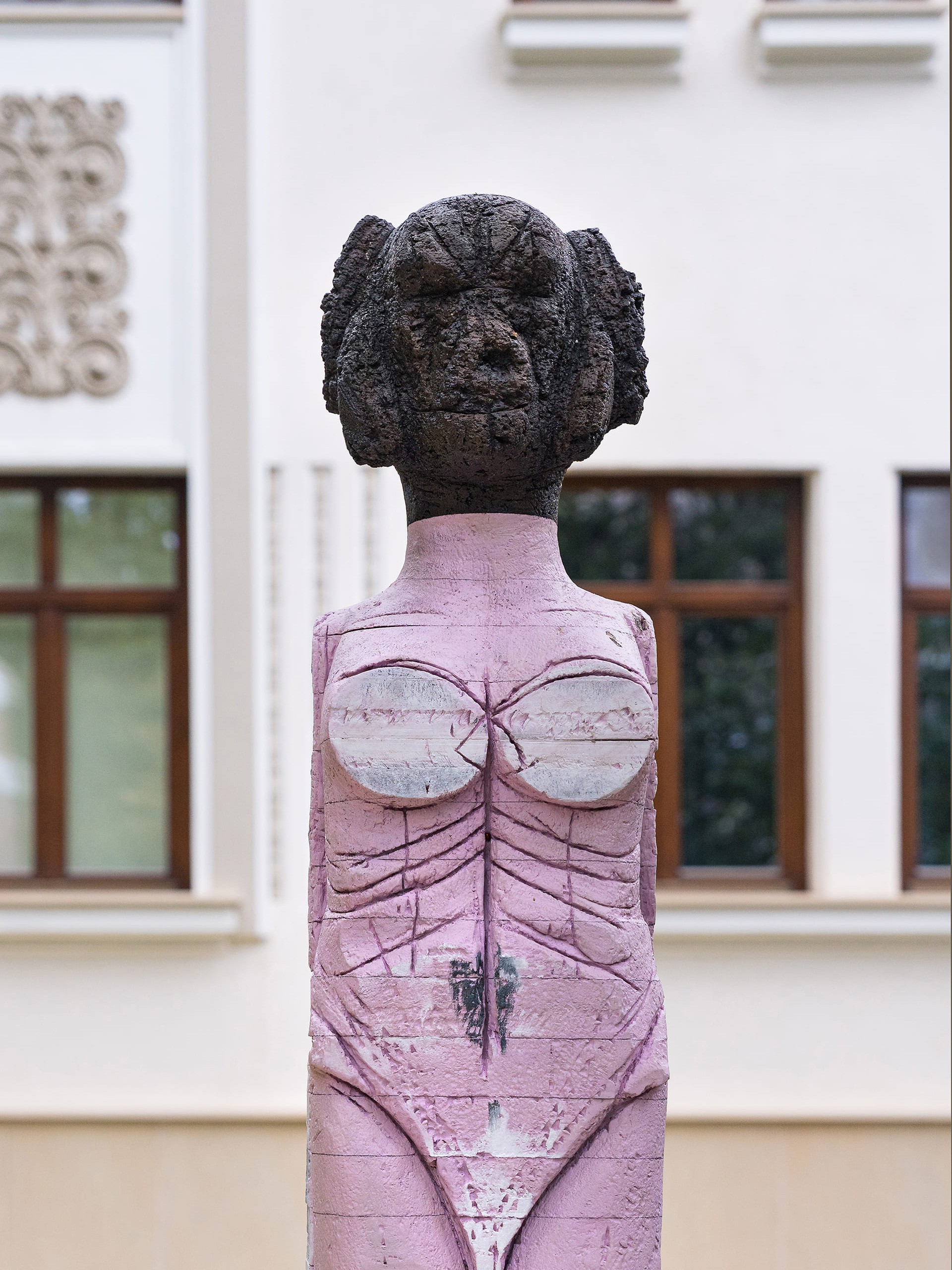 Outdoor sculpture of a stylized human figure with a rough, dark textured head and lower legs. The torso and upper legs are painted a muted pink, with carved linear markings suggesting anatomical features. The chest area features two rounded white forms, and the surface shows signs of weathering and cracks throughout.