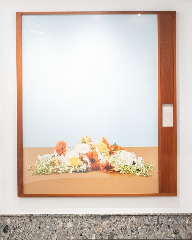 A large photograph framed in wood, depicting a horizontal arrangement of orange, yellow, and white flowers laid on a flat brown surface. The pale blue background creates a serene, minimalist composition that evokes stillness and contemplation.