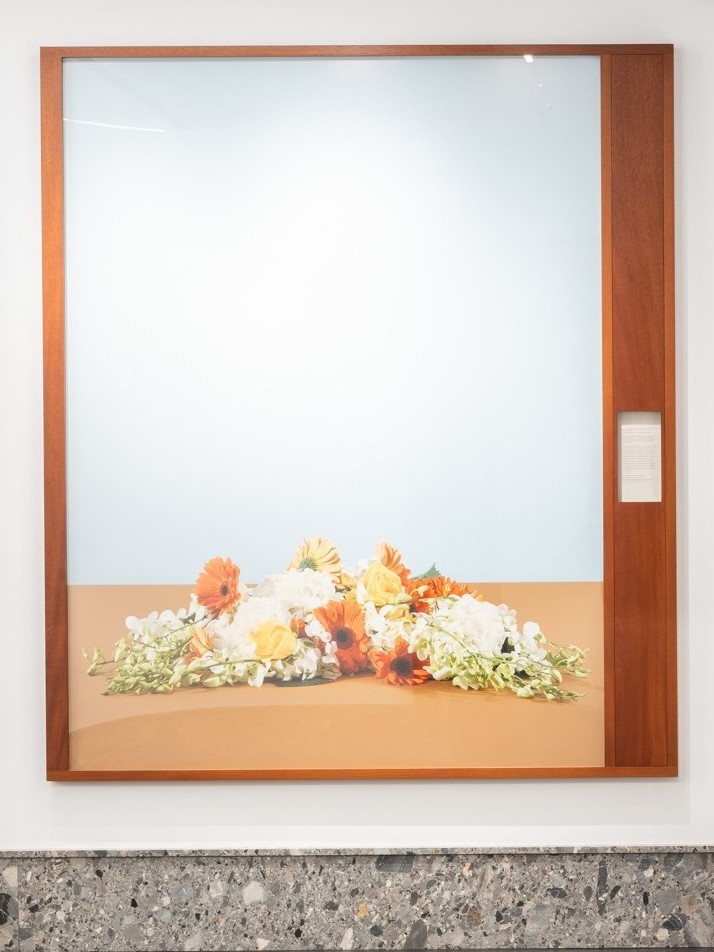A large photograph framed in wood, depicting a horizontal arrangement of orange, yellow, and white flowers laid on a flat brown surface. The pale blue background creates a serene, minimalist composition that evokes stillness and contemplation.
