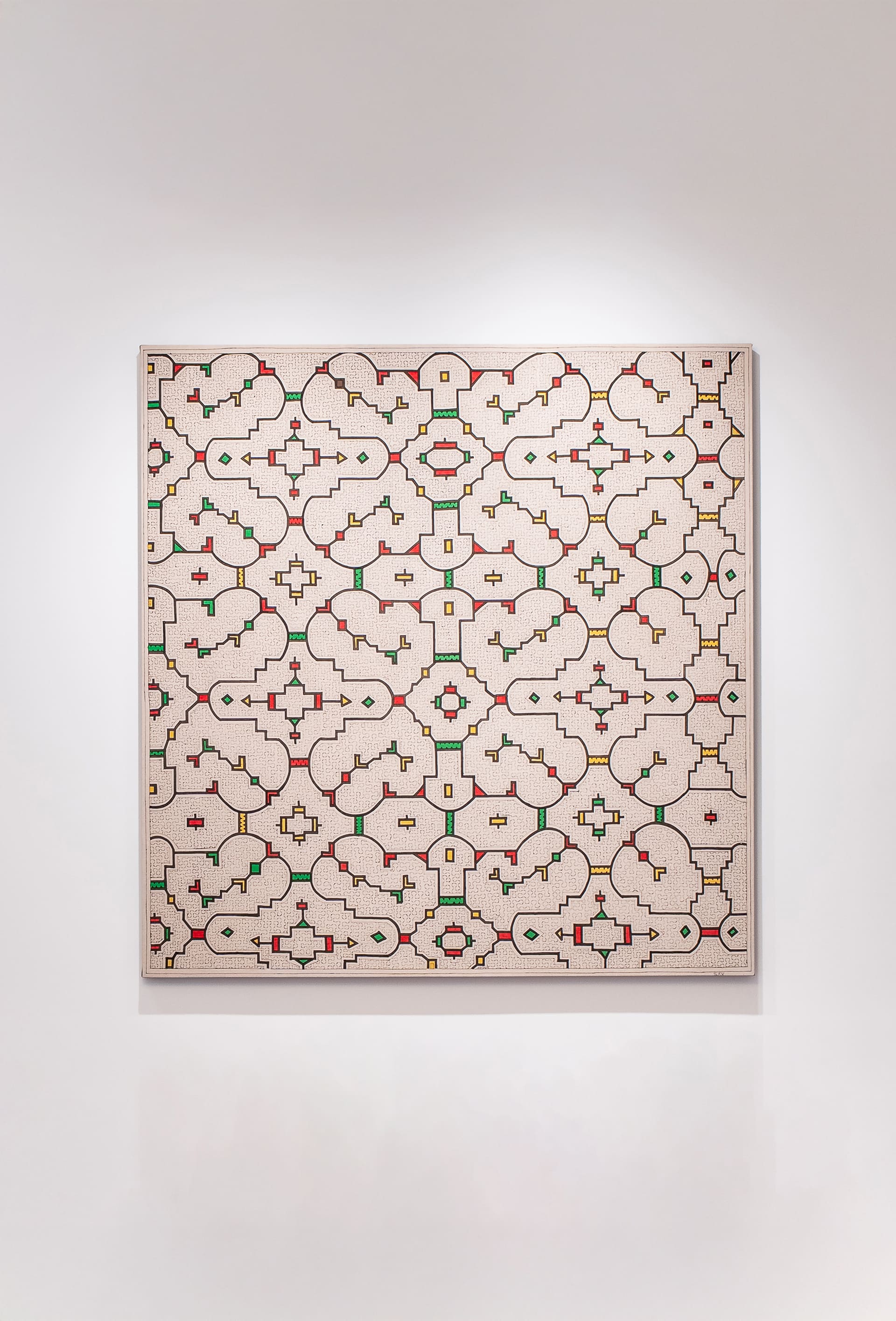 A large geometric artwork hangs on a white wall, featuring a repeating pattern of interconnected shapes resembling stylized tiles or architectural motifs. The design is composed of fine black lines forming symmetrical loops, arches, and cross-like figures, accented with small blocks of red, green, and yellow. The background is a warm, textured beige that gives the piece a hand-drawn, intricate quality, evoking the look of an ornamental lattice.
