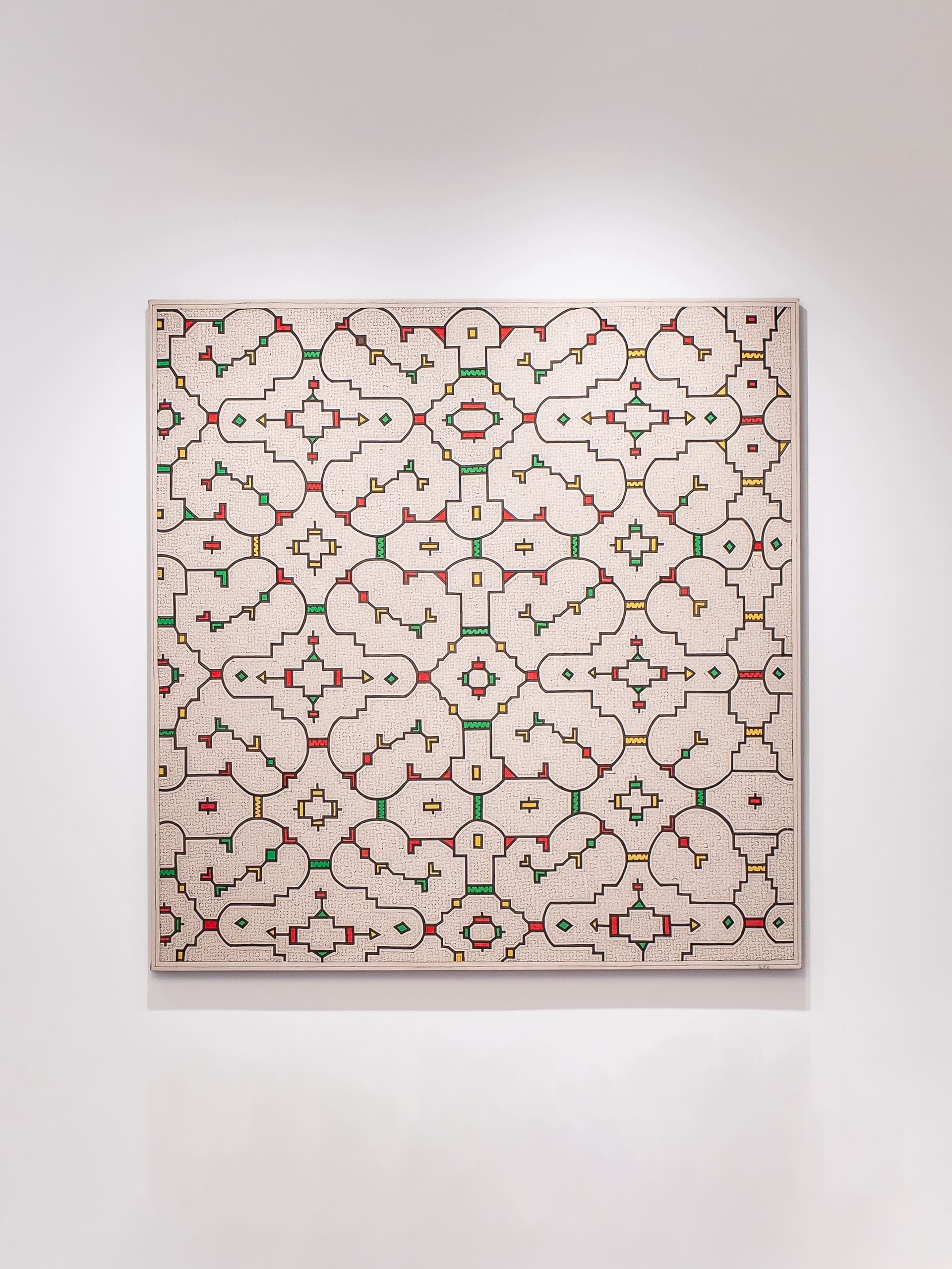 A large geometric artwork hangs on a white wall, featuring a repeating pattern of interconnected shapes resembling stylized tiles or architectural motifs. The design is composed of fine black lines forming symmetrical loops, arches, and cross-like figures, accented with small blocks of red, green, and yellow. The background is a warm, textured beige that gives the piece a hand-drawn, intricate quality, evoking the look of an ornamental lattice.