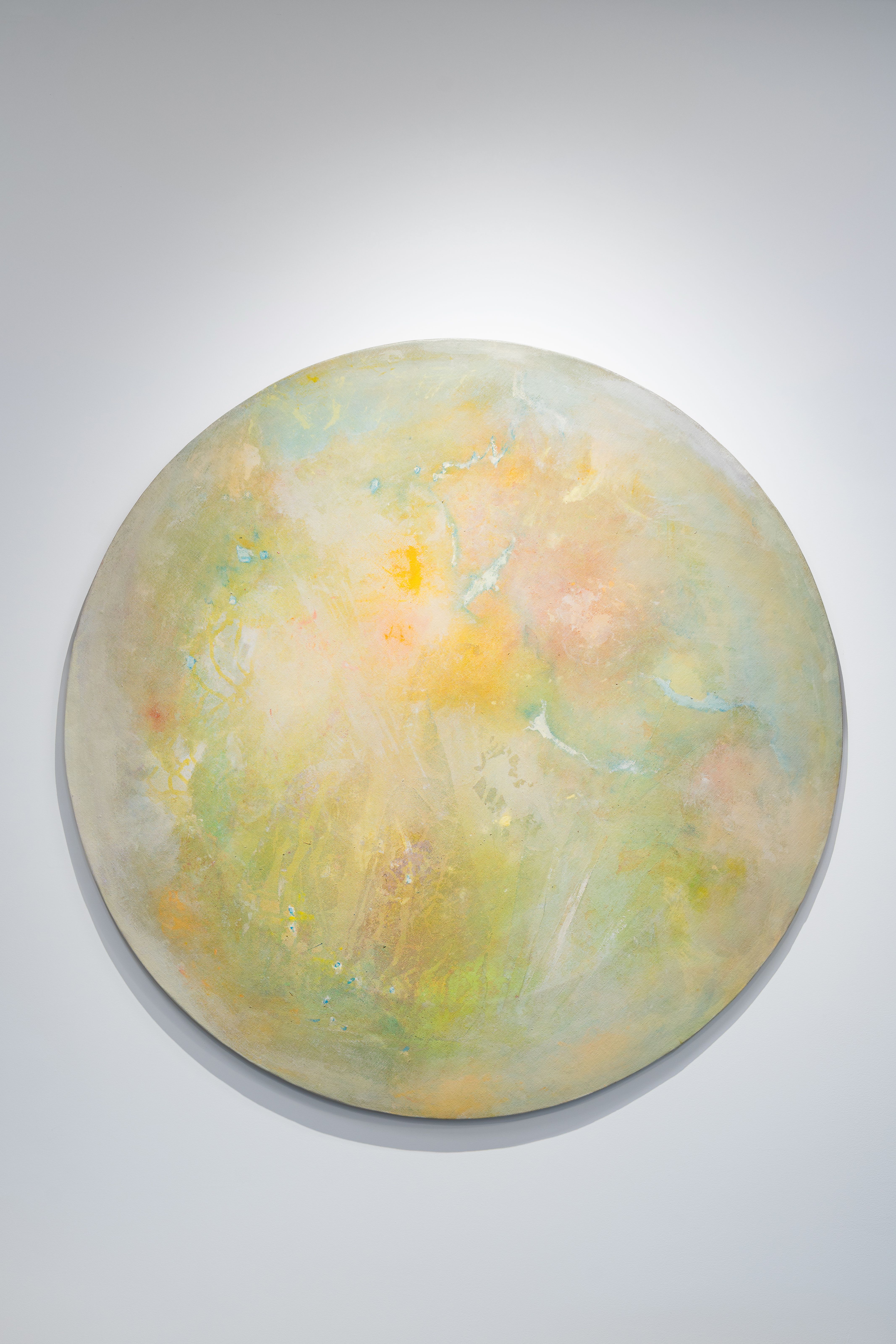 A circular abstract painting in soft tones of yellow, green, and peach. The surface has a hazy, atmospheric quality, resembling a celestial body or watercolor planet glowing gently against a white wall.