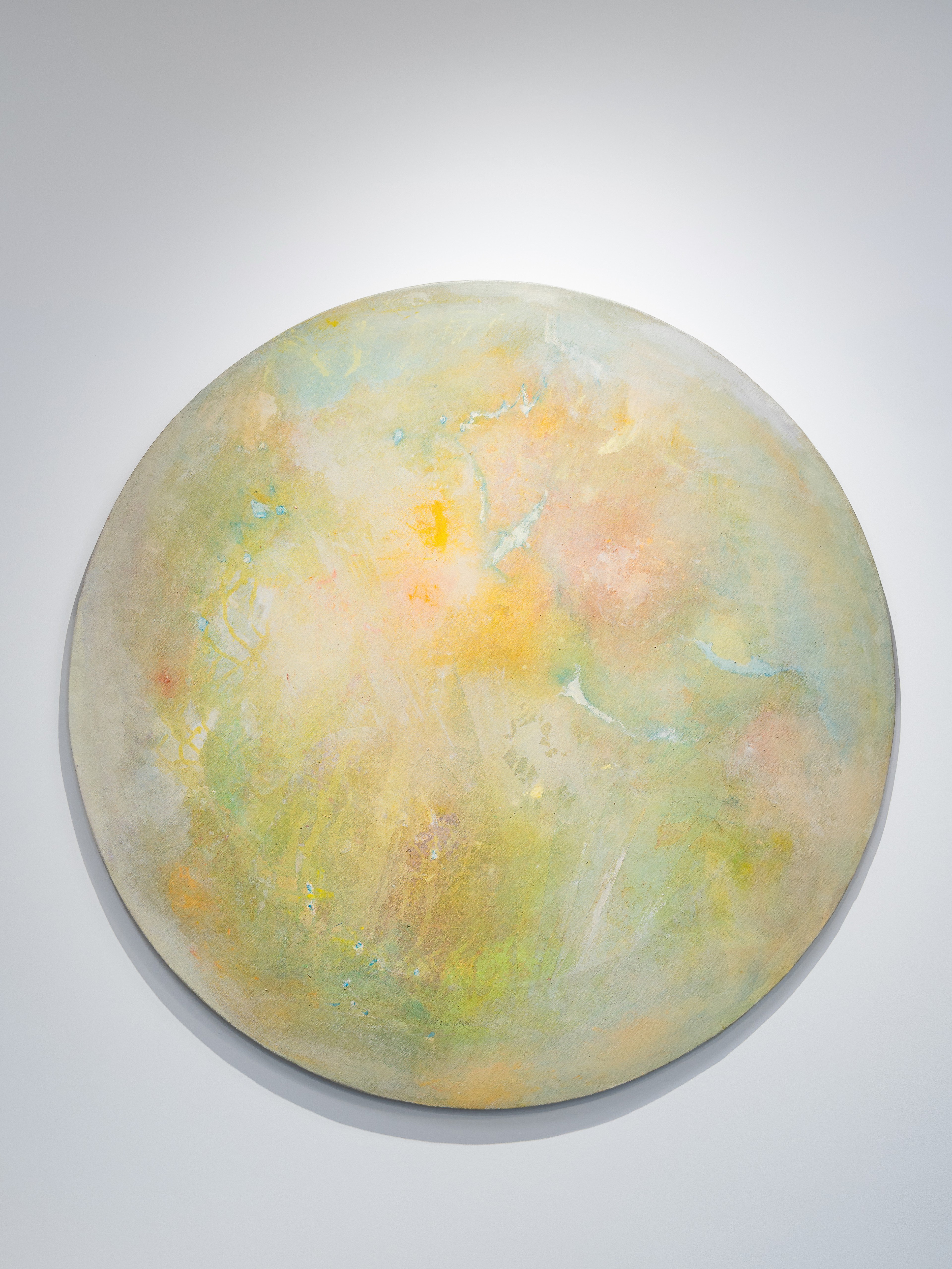 A circular abstract painting in soft tones of yellow, green, and peach. The surface has a hazy, atmospheric quality, resembling a celestial body or watercolor planet glowing gently against a white wall.