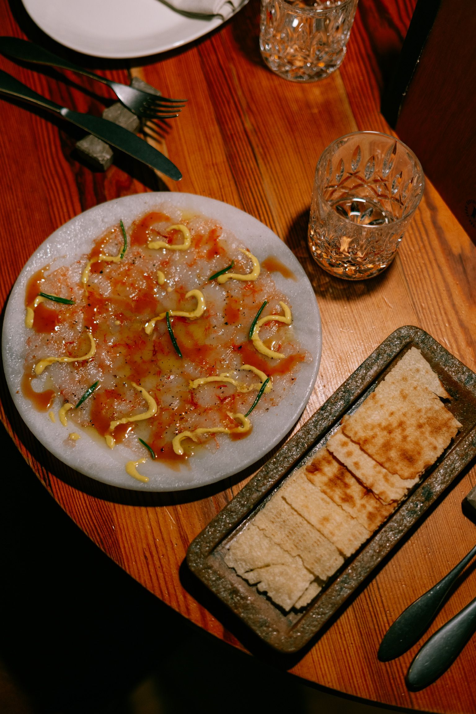 Láminas finas de pescado aliñadas en un plato de mármol, con crujiente de pan y vasos de agua