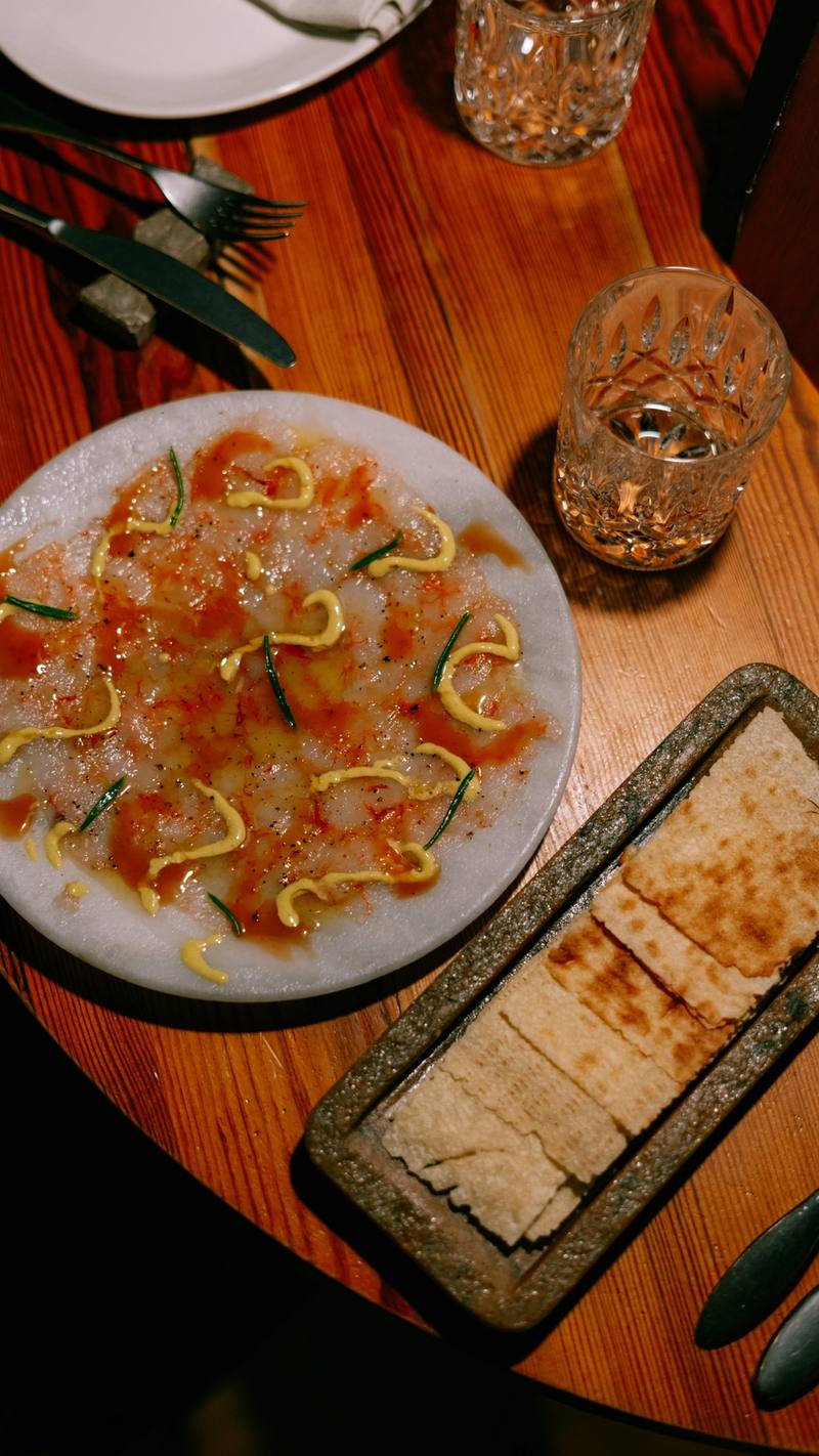 Láminas finas de pescado aliñadas en un plato de mármol, con crujiente de pan y vasos de agua