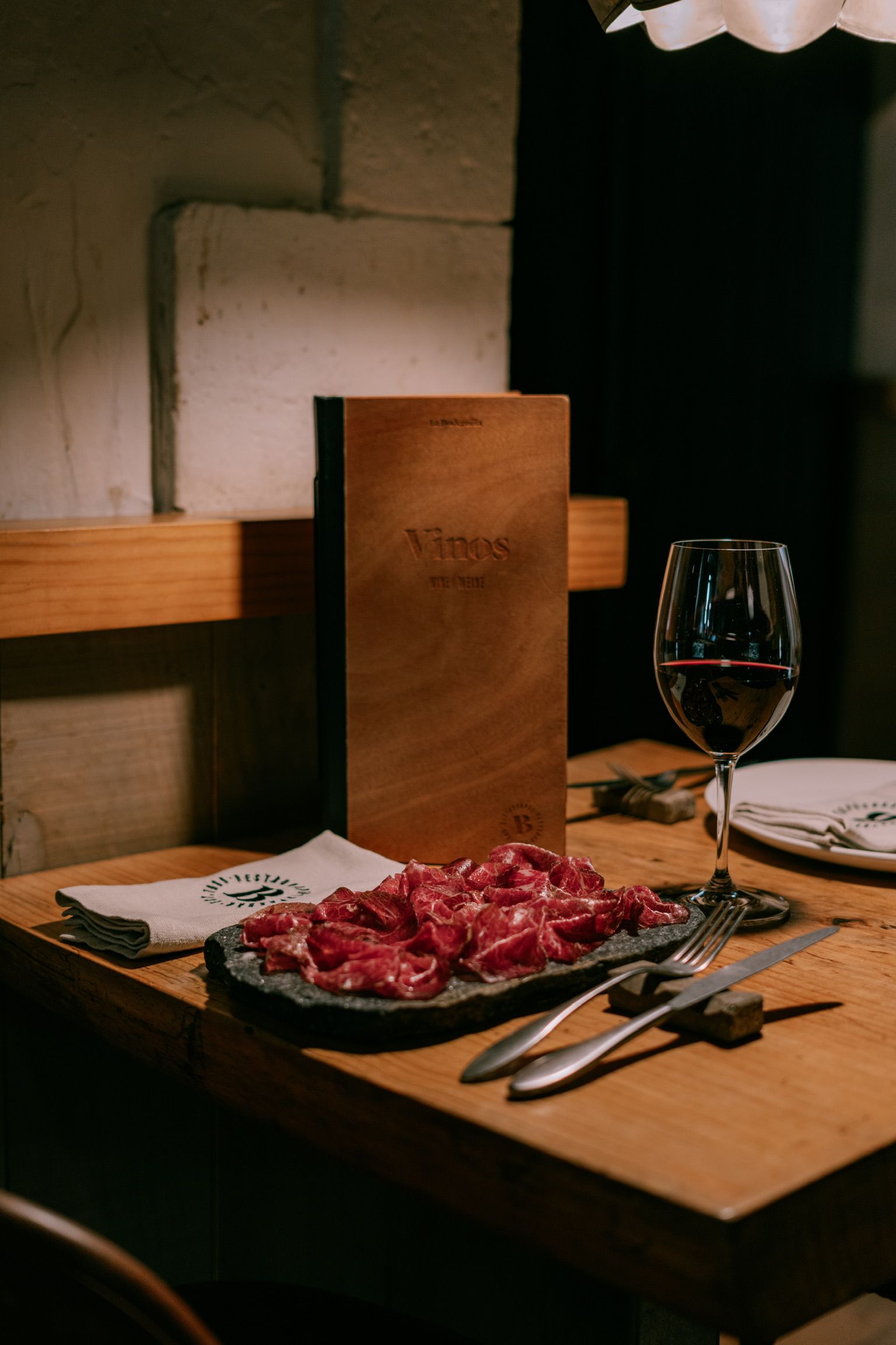 Plato de embutido en láminas sobre piedra, copa de vino tinto y carta de vinos en una mesa de madera