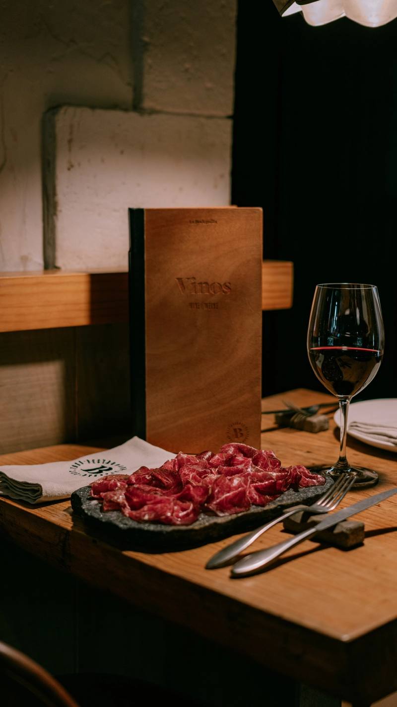 Plato de embutido en láminas sobre piedra, copa de vino tinto y carta de vinos en una mesa de madera