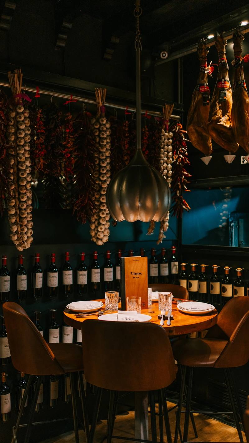 Mesa alta junto a una pared de botellas de vino, con jamones, ristras de ajos y guindillas colgados en La Bodeguilla