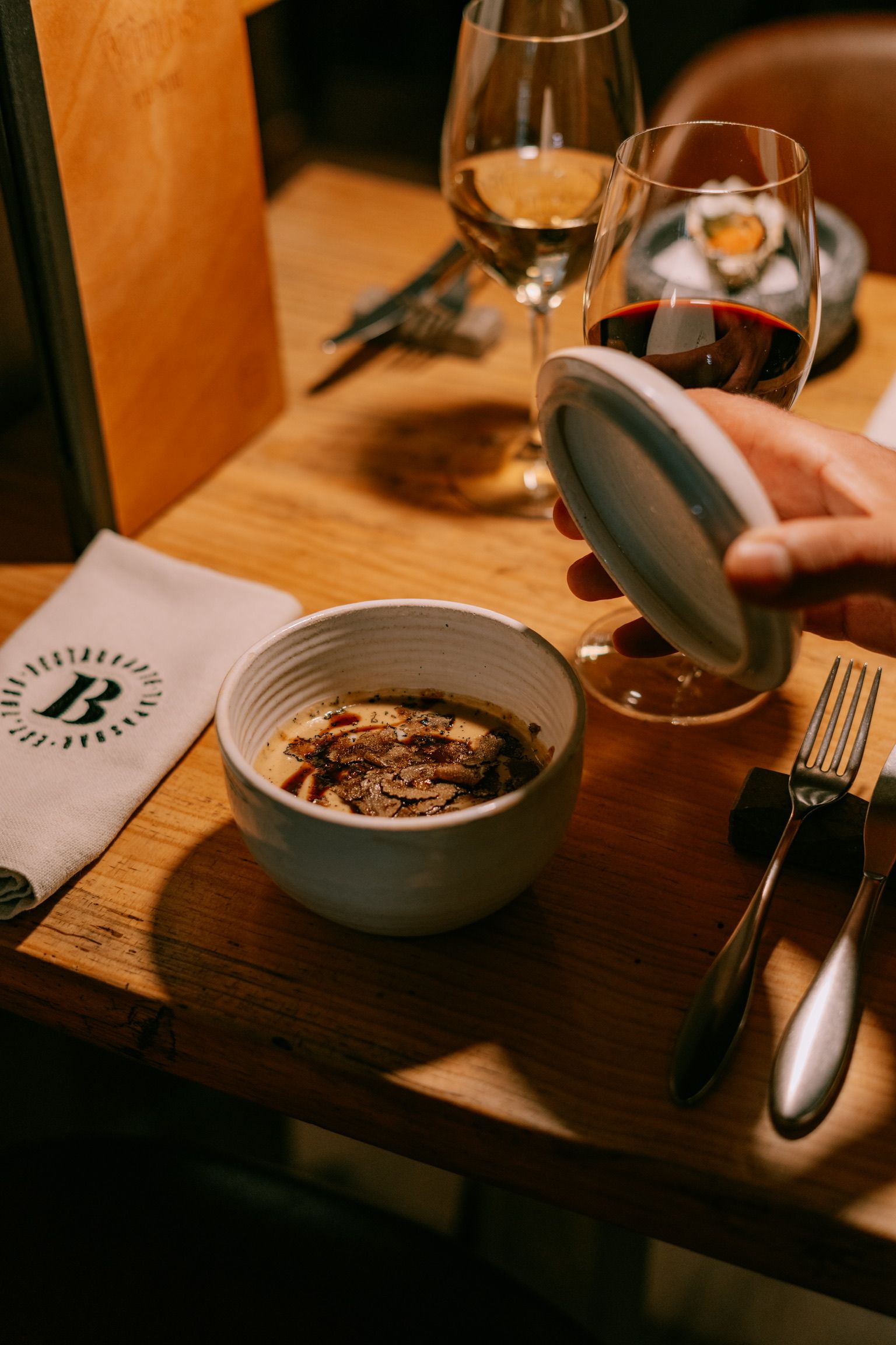Cuenco de crema con láminas de trufa negra, destapado en la mesa junto a dos copas de vino