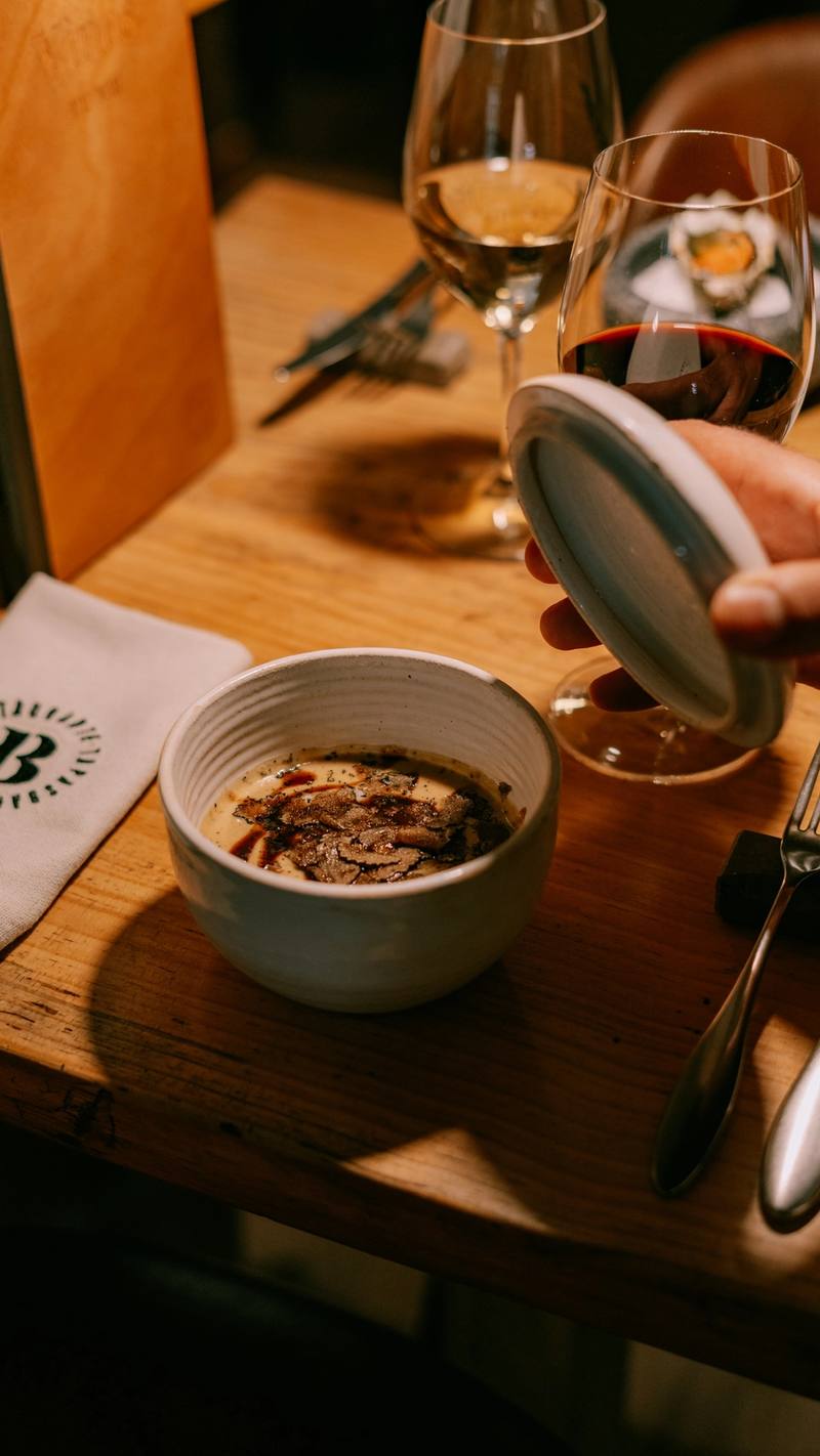 Cuenco de crema con láminas de trufa negra, destapado en la mesa junto a dos copas de vino