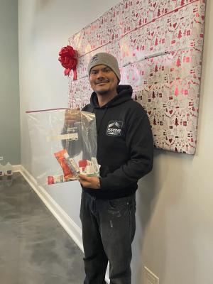 A RoofingSource teammate holding a bagged gift beside a wrapped and bowed board at a holiday gift drive.