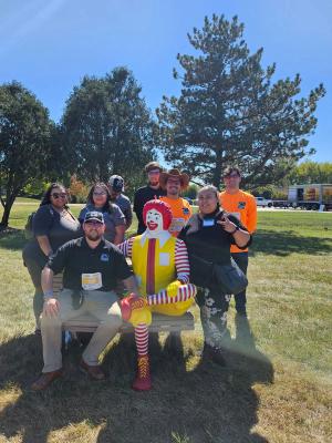 Eight teammates gathered on the grass around a life-size Ronald McDonald bench statue at a charity fun run, two of them wearing race bibs.