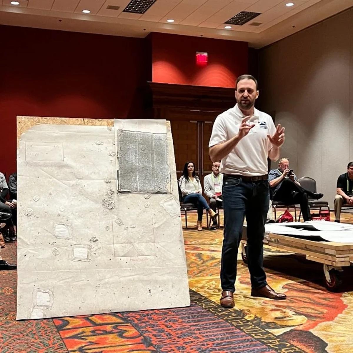 A RoofingSource presenter speaking beside a roof-section sample at RFMA