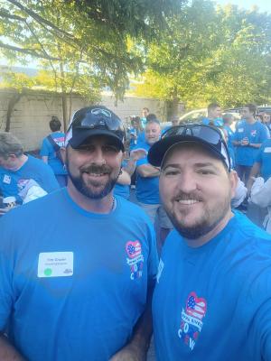 Two RoofingSource teammates in matching charity volunteer-day shirts and company caps, with a crowd of volunteers in the same shirts behind them.