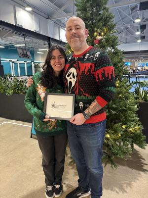 Two teammates in holiday sweaters holding a framed Ronald McDonald House Charities certificate beside a decorated Christmas tree.