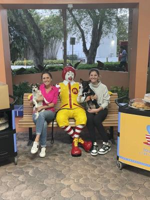 Two teammates each holding a small dog on a bench beside a life-size Ronald McDonald statue, with meal carts either side.