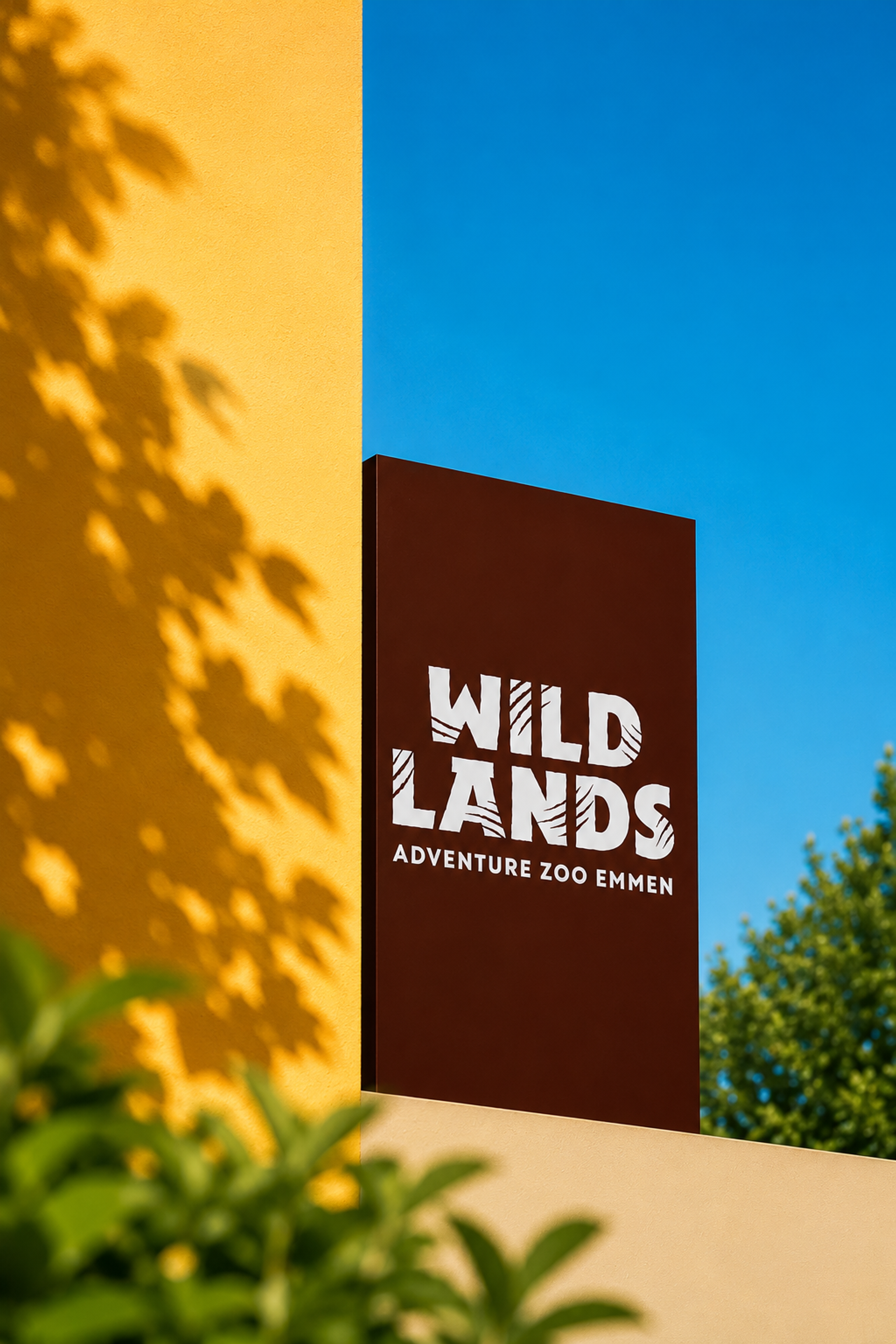Wildlands Adventure Zoo Emmen-logo tegen een heldere blauwe lucht met warme kleuren en groene beplanting op de voorgrond.