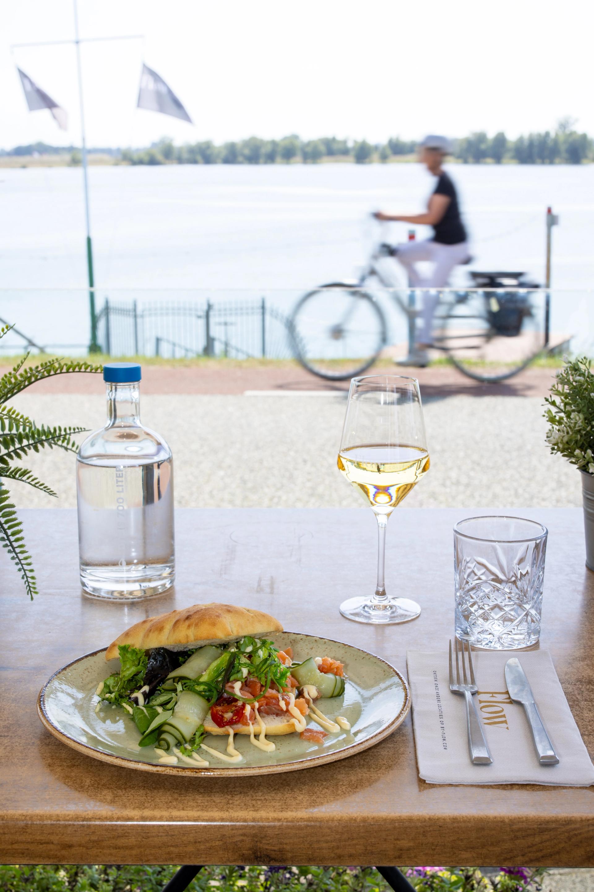 Lunchgerecht en een glas witte wijn op het terras aan de Maas, met een fietser op de achtergrond