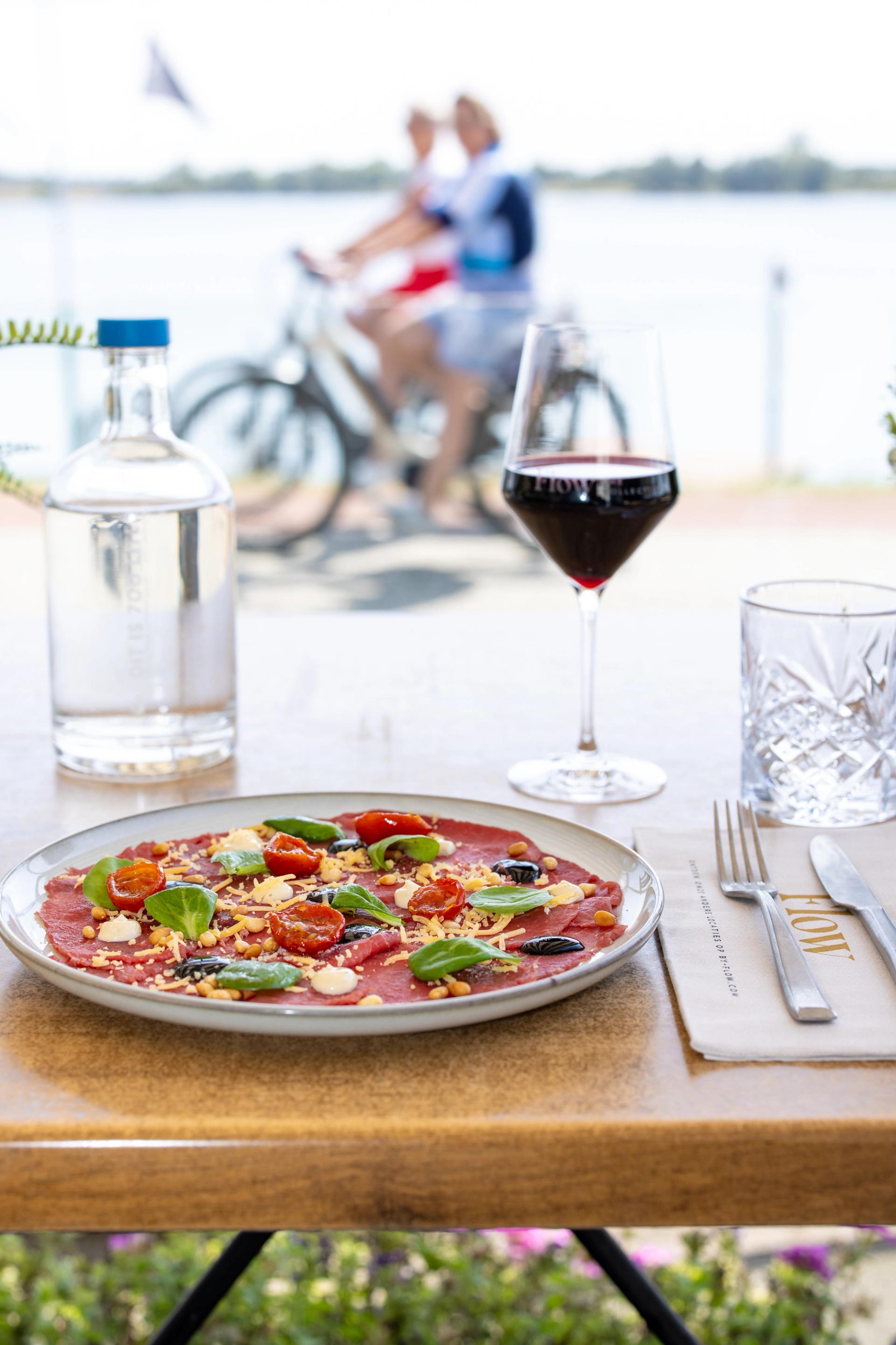 carpaccio gerecht en glas rode wijn op opgedekte tafel met uitzicht op de maas en twee fietsers rijden langs