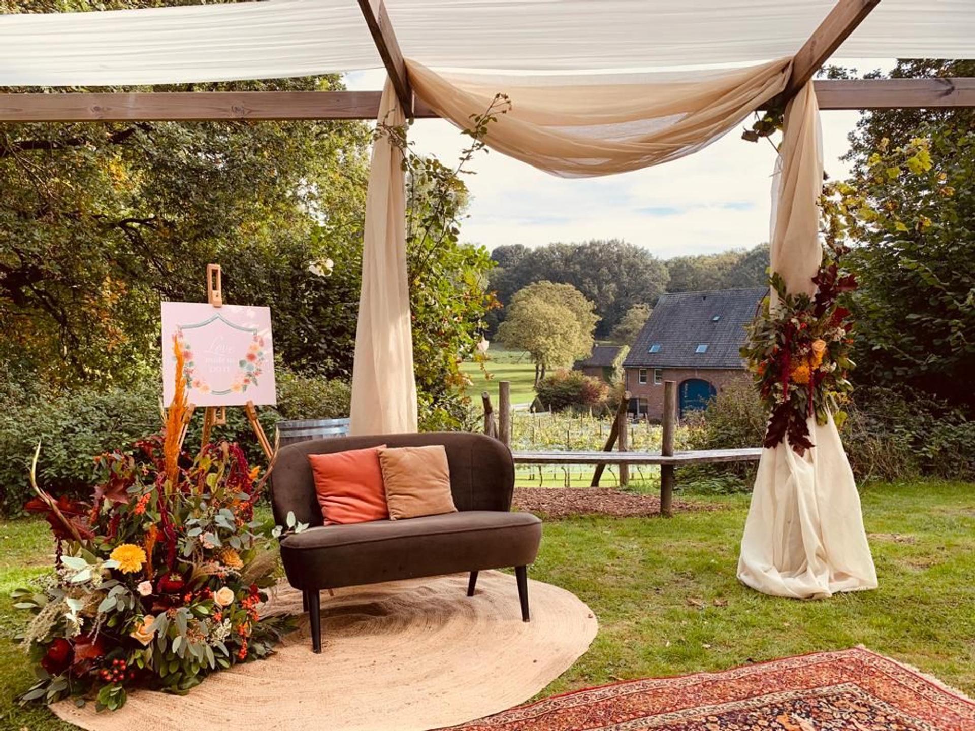 Outdoor wedding at Winselerhof in Limburg with an atmospherically dressed ceremony set-up under a green pergola, white chairs on the lawn and a romantic pink loveseat