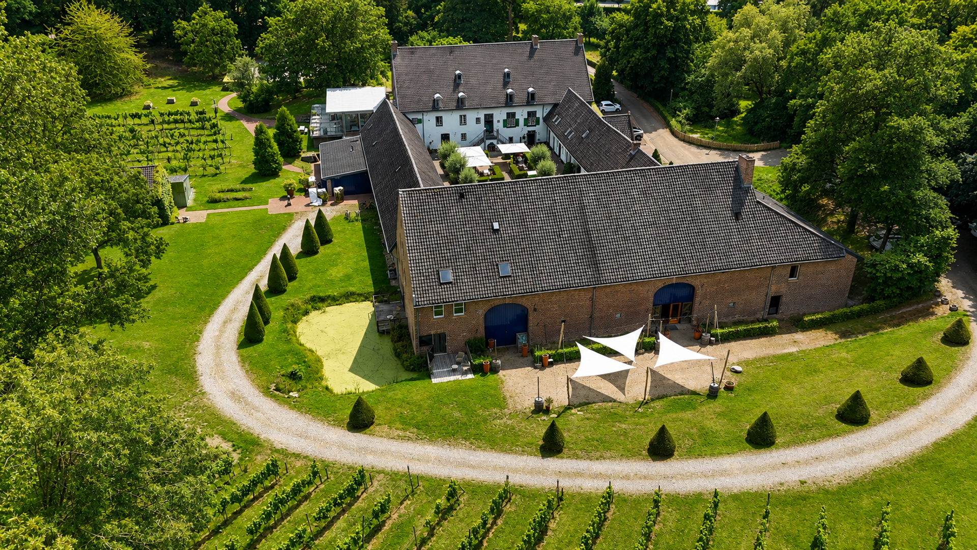Een luchtfoto van Hotel Winselerhof in Zuid-Limburg. Het historische hotelcomplex wordt omringd door groene gazons, bomen en wijngaarden. De gebouwen liggen rond een binnenplaats en worden verbonden door wandelpaden. Op de voorgrond zijn rijen wijnstokken zichtbaar, terwijl het landgoed zich uitstrekt in een rustige, natuurlijke omgeving.