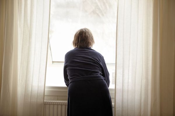 An elderly person stands alone at a window, seen from behind, wearing a dark robe. Soft light filters through sheer white curtains.