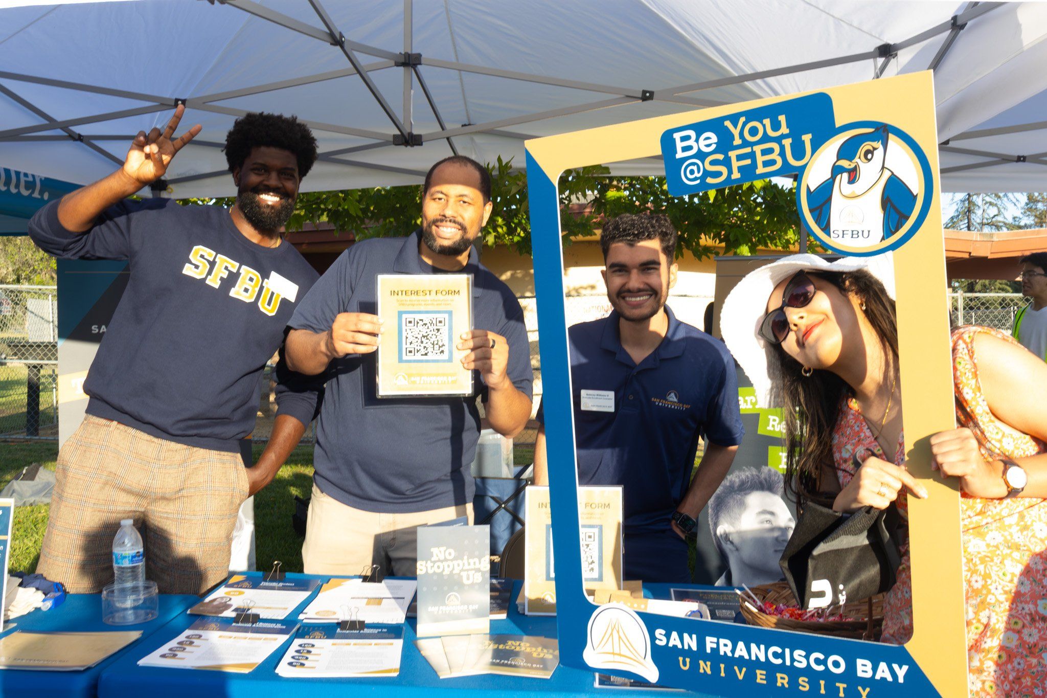 Three SFBU staff members at an outdoor recruiting table under a canopy, holding up a QR-code interest form and a "Be You @SFBU" photo frame
