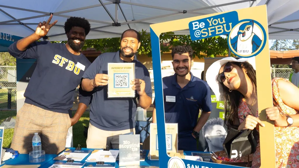 Three SFBU staff members at an outdoor recruiting table under a canopy, holding up a QR-code interest form and a "Be You @SFBU" photo frame