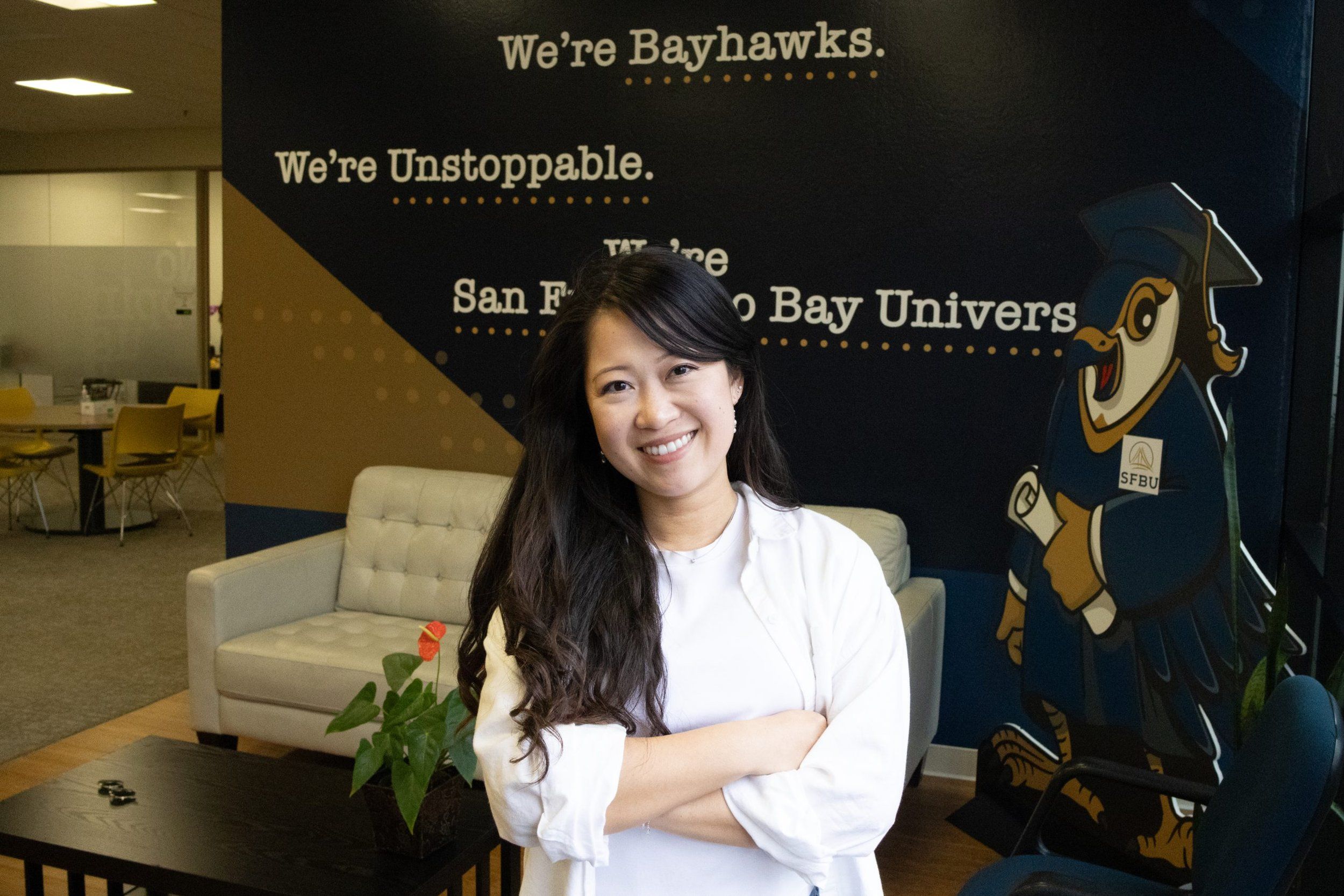 An SFBU staff member standing with arms folded in a campus lounge in front of a wall mural reading "We're Bayhawks. We're Unstoppable." beside the university's hawk mascot