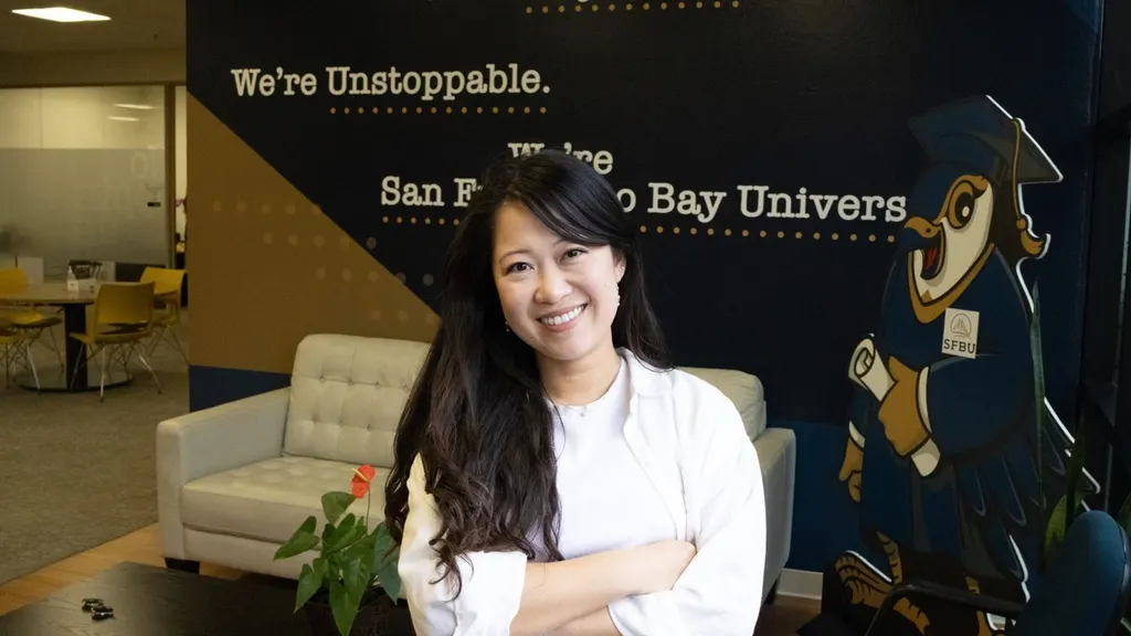 An SFBU staff member standing with arms folded in a campus lounge in front of a wall mural reading "We're Bayhawks. We're Unstoppable." beside the university's hawk mascot