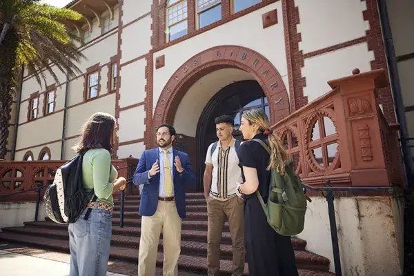 Three Flagler College students with backpacks talking with a faculty member on the steps of the campus library