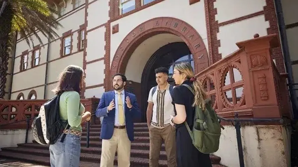 Three Flagler College students with backpacks talking with a faculty member on the steps of the campus library