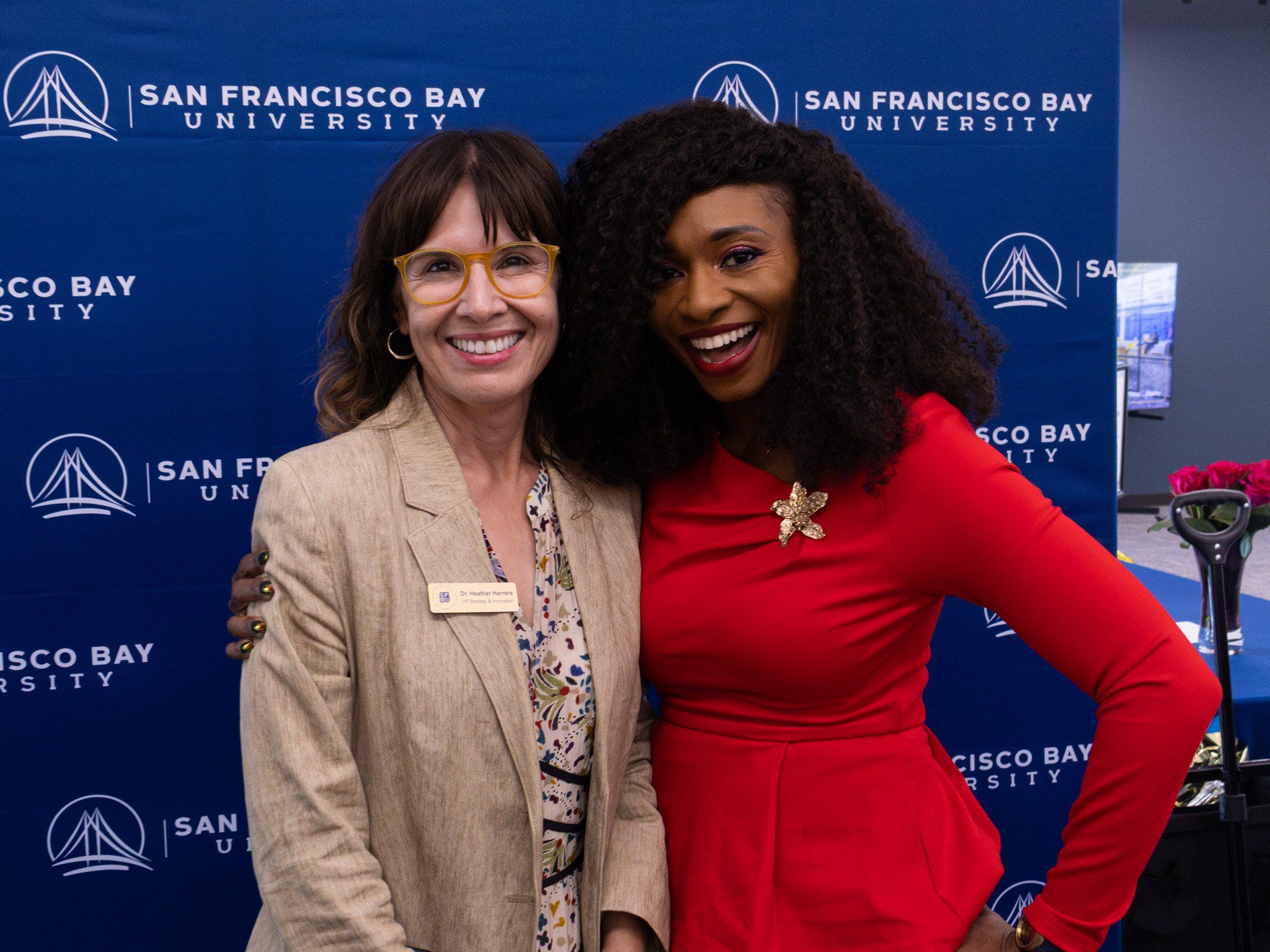 Two women smiling arm in arm in front of a San Francisco Bay University step-and-repeat banner at a campus kickoff event