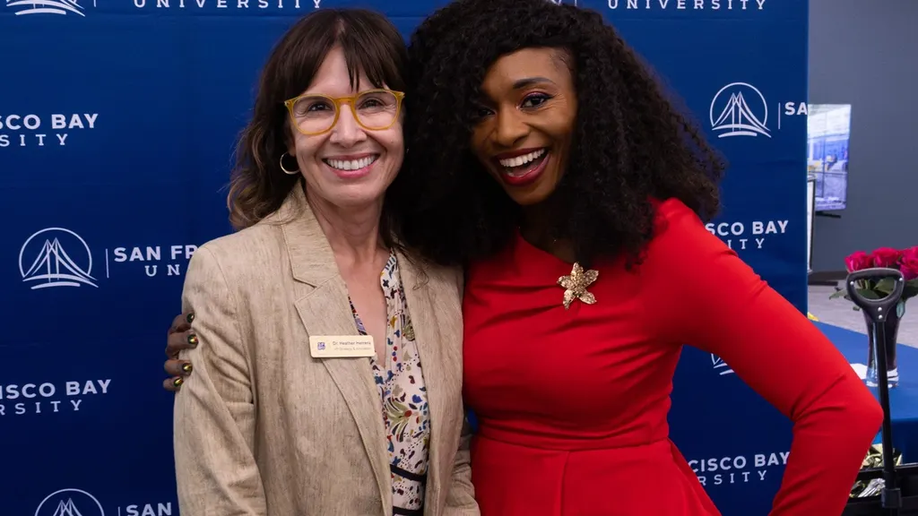 Two women smiling arm in arm in front of a San Francisco Bay University step-and-repeat banner at a campus kickoff event