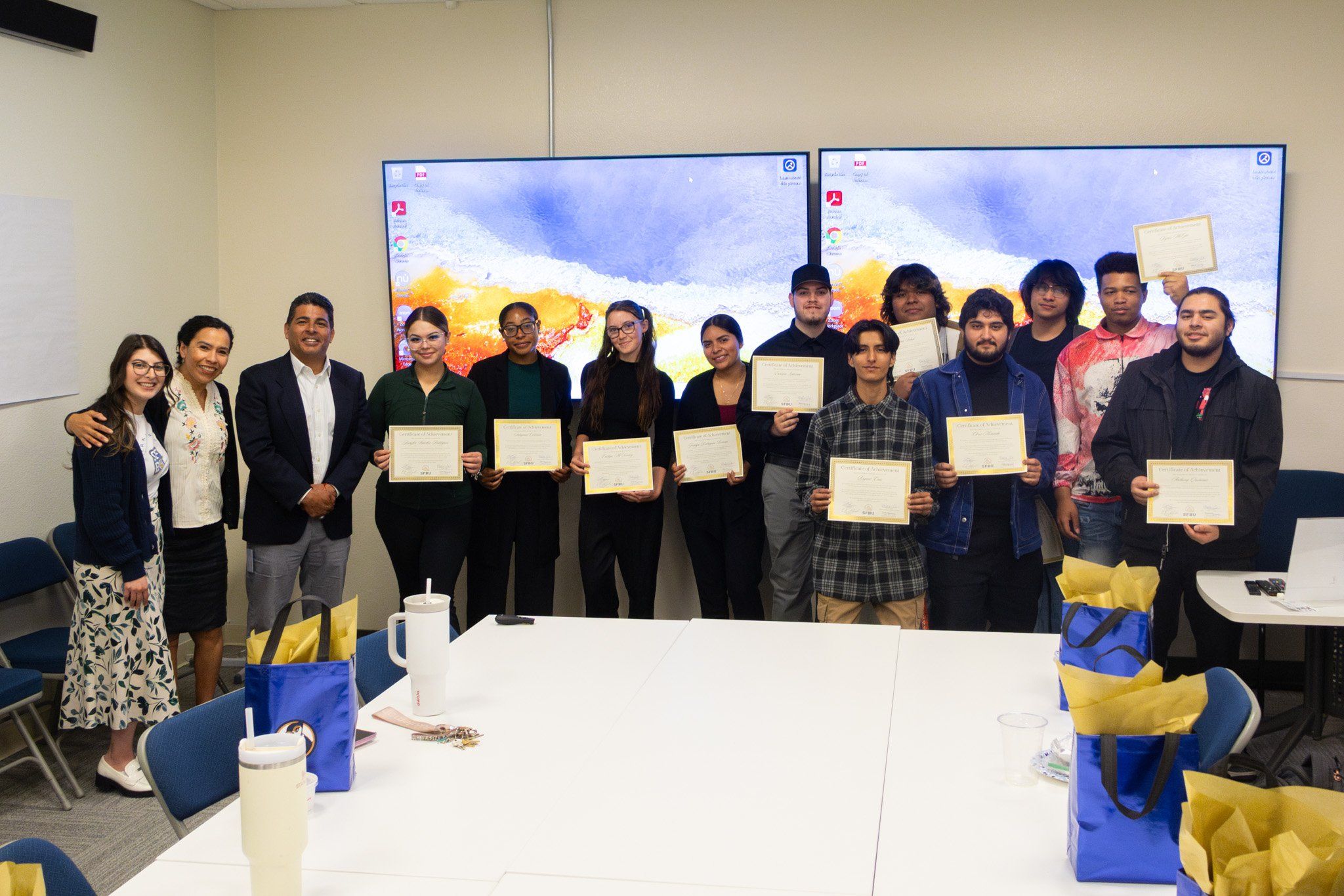 A group of SFBU students standing with framed certificates alongside faculty in a classroom, with presentation screens behind them