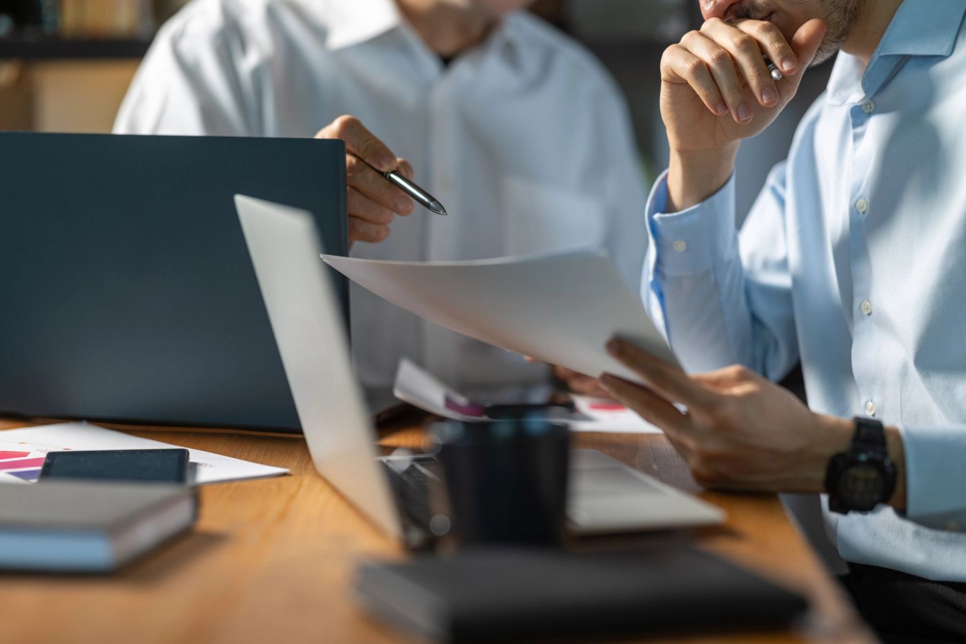Two colleagues in shirts reviewing printed documents beside a laptop at a desk.