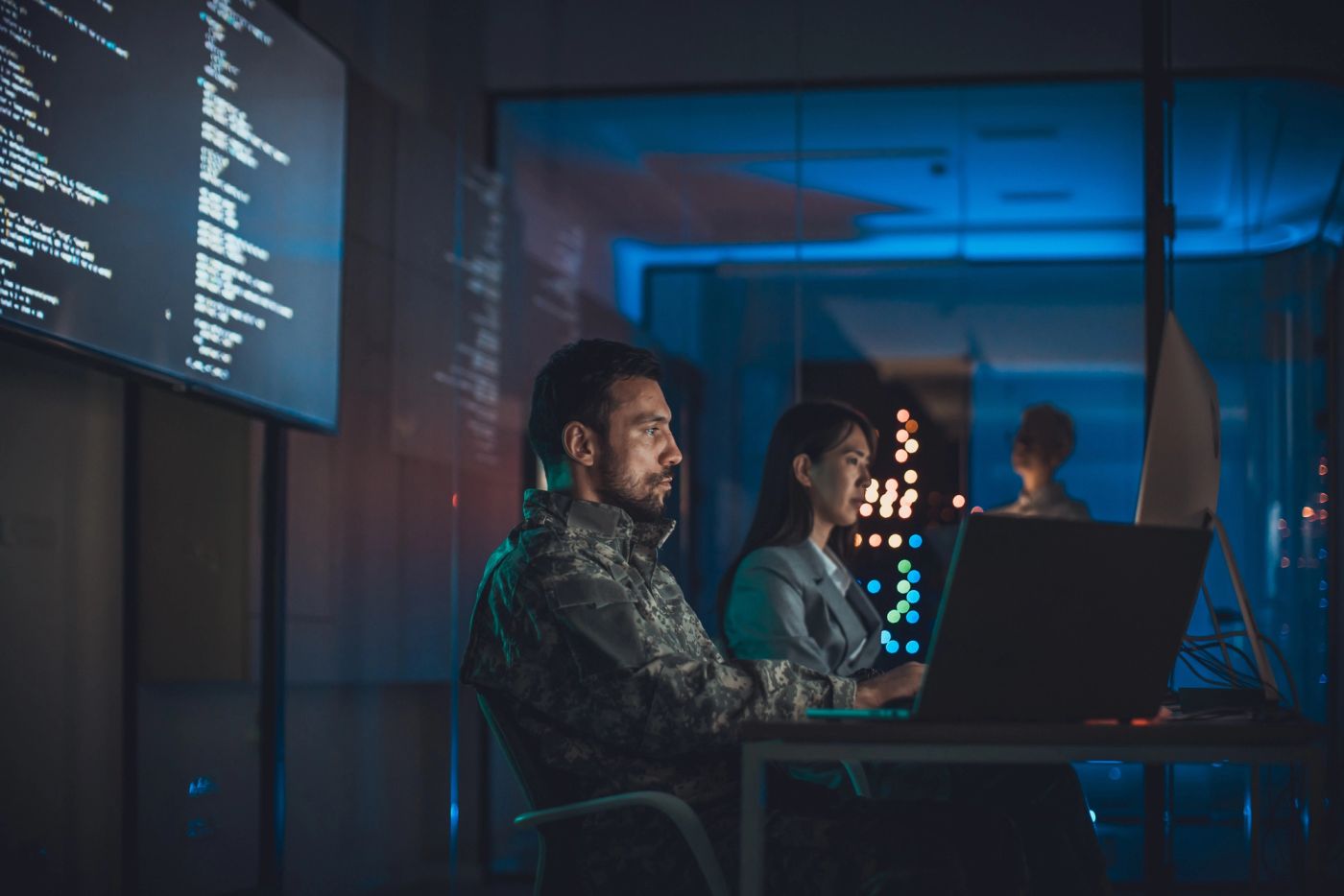 Two analysts, one in military fatigues, working at a laptop in a dim control room with code on a wall display.