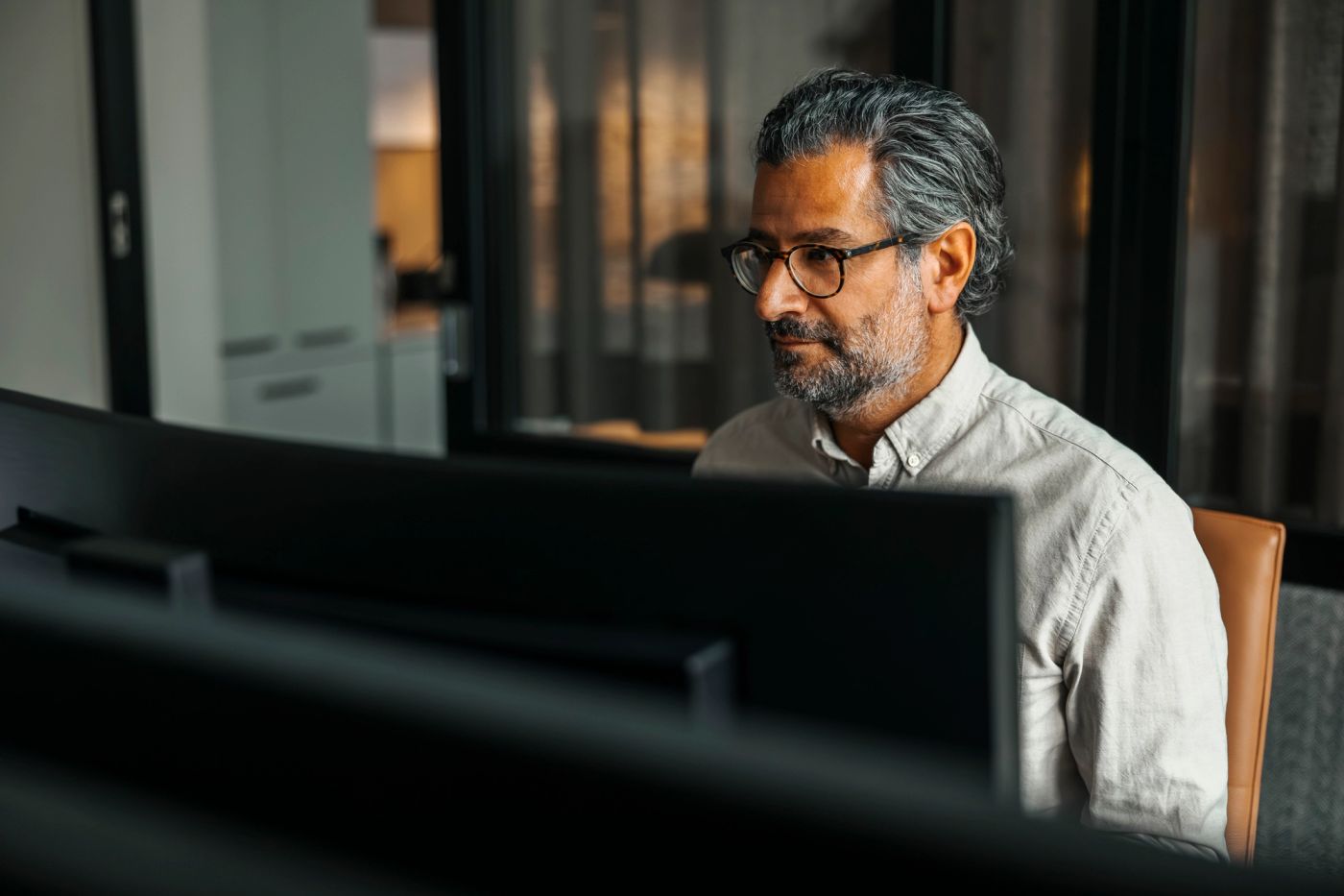 A man with glasses and grey hair seated at a desk in front of computer monitors.